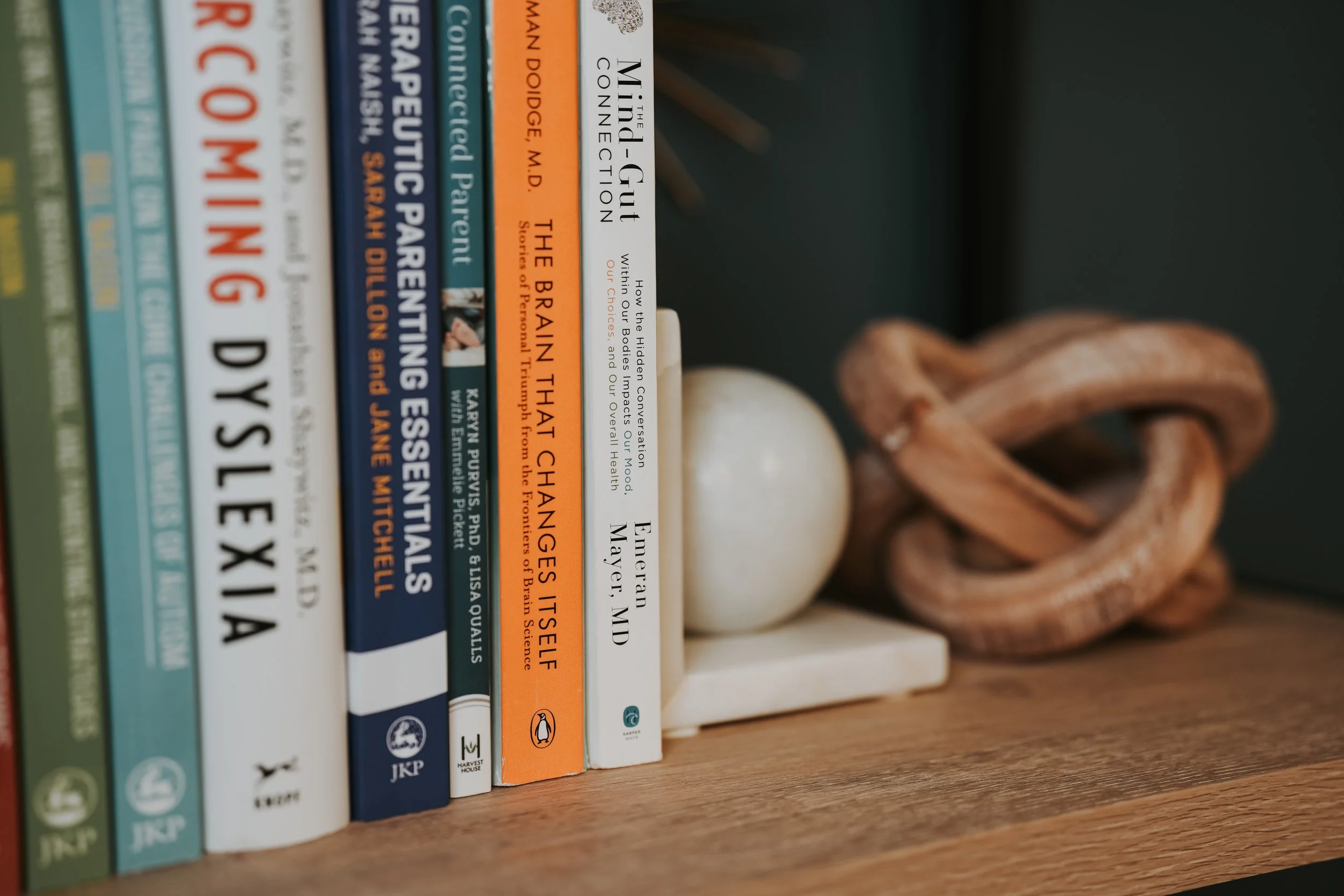 A row of books on a wooden shelf, with titles related to health, psychology, and self-help, next to a decorative wooden knot and a white spherical object.