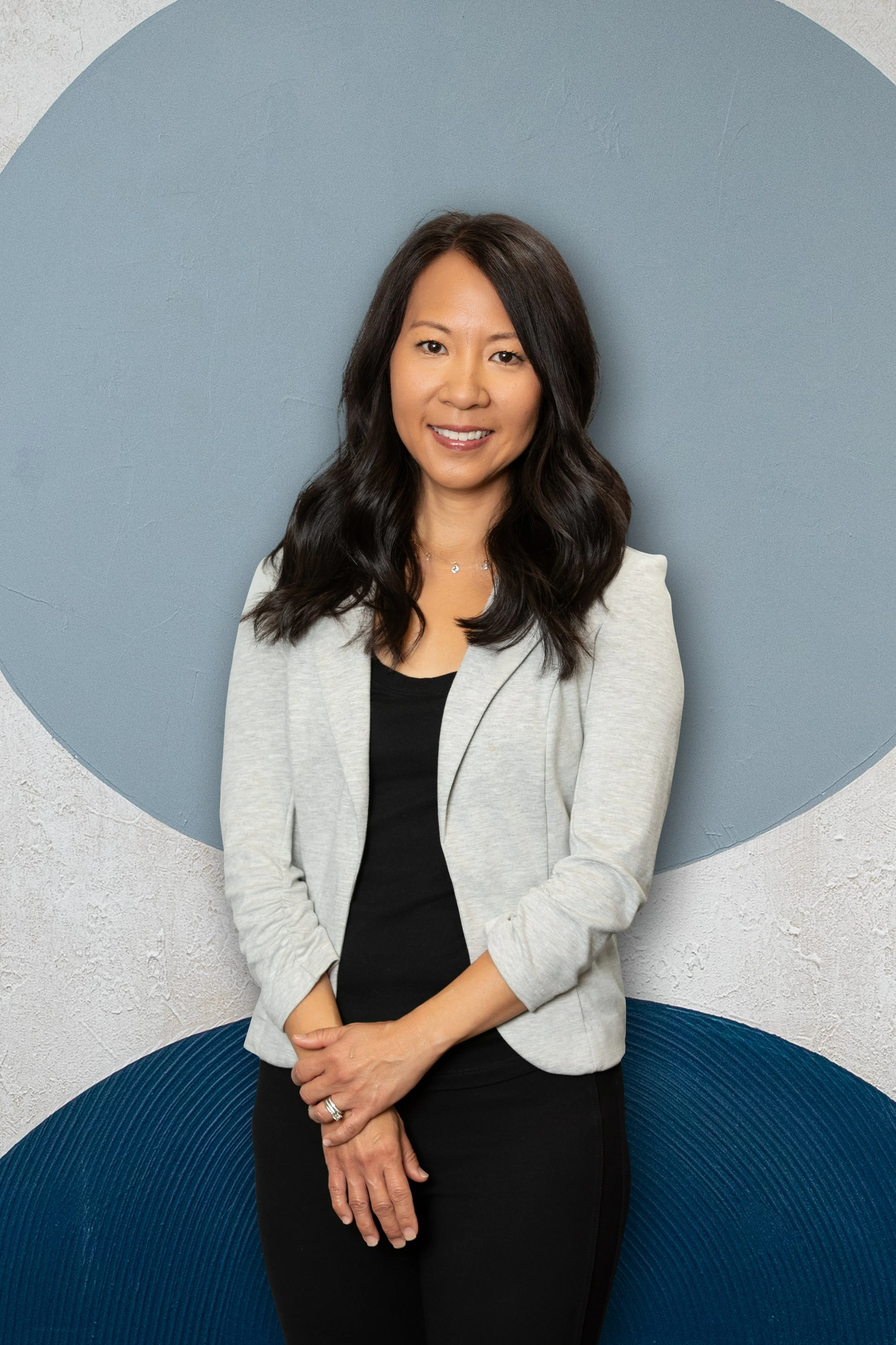 A woman with long dark hair wearing a light gray blazer over a black top, standing against a background with blue and white textured walls.