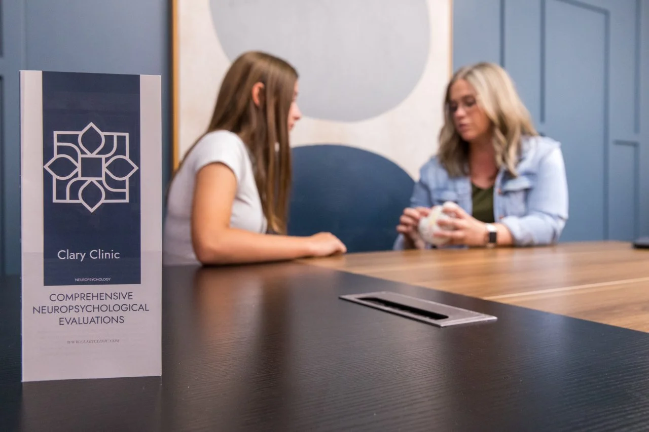 A woman and a girl sitting at a table in a clinic, discussing. A sign on the table reads 'Clary Clinic, Comprehensive Neuropsychological Evaluations'.