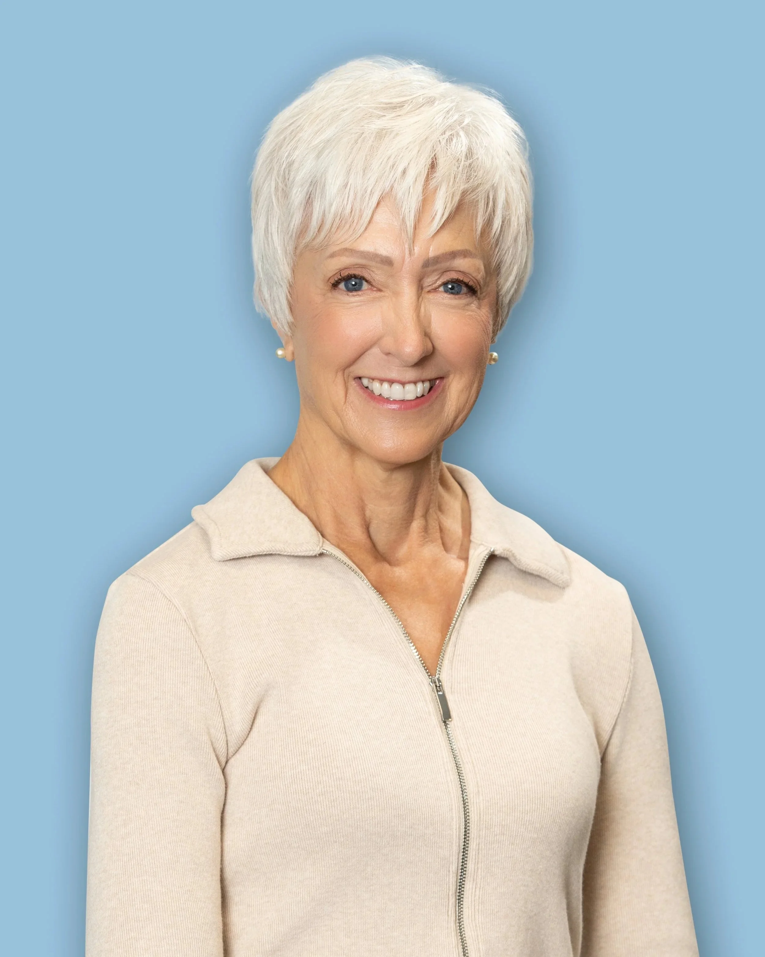 Portrait of a senior woman with short white hair, smiling, wearing a beige zip-up sweater, against a light blue background.
