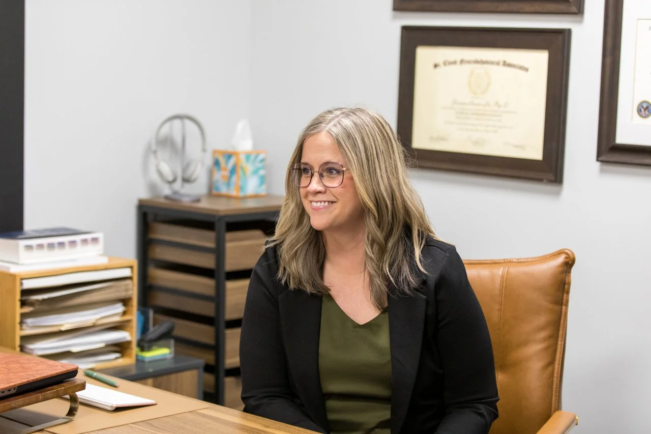 A woman with shoulder-length blonde hair, glasses, and a black blazer sits in a chair in an office, smiling. The office has framed certificates on the wall, a wooden desk, and shelves with papers and office supplies.