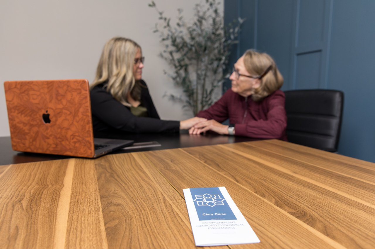 Two women sitting at a wooden table, holding hands and talking, with a laptop and a pamphlet on the table.