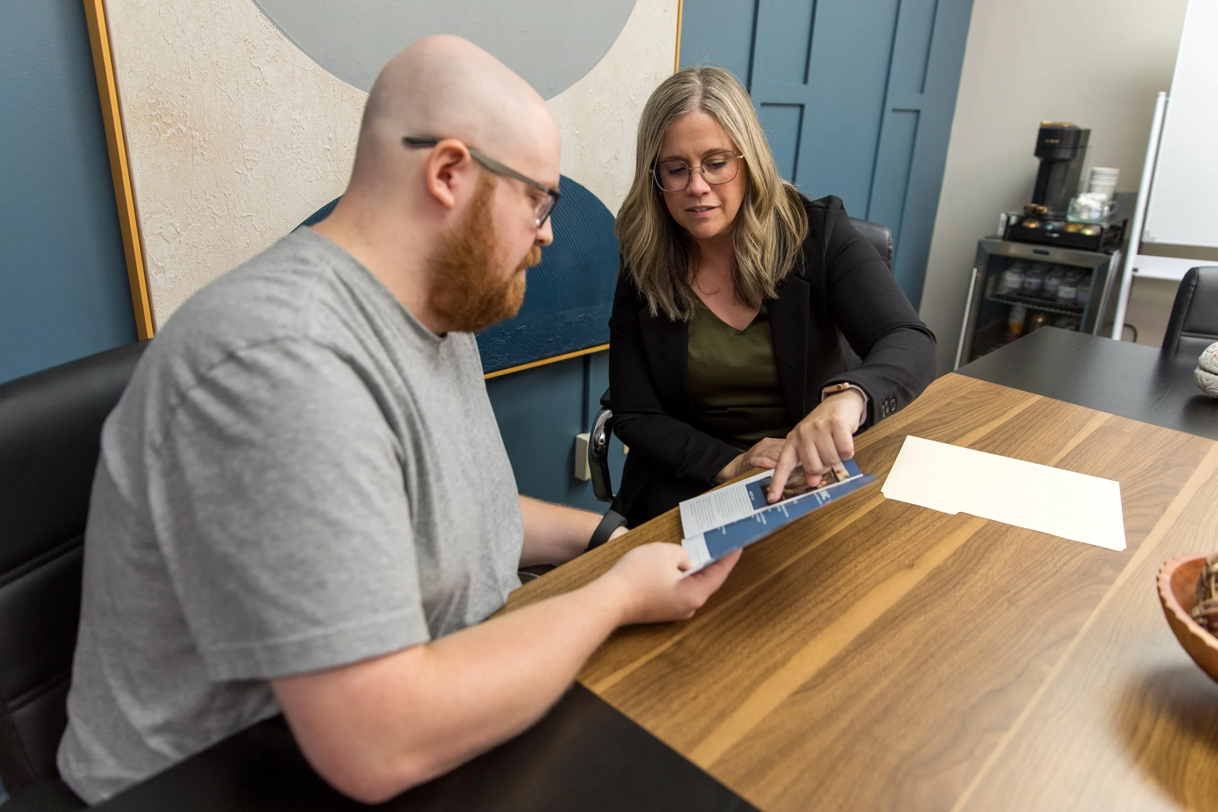 A woman showing a man something on a brochure or document at a meeting table in an office.
