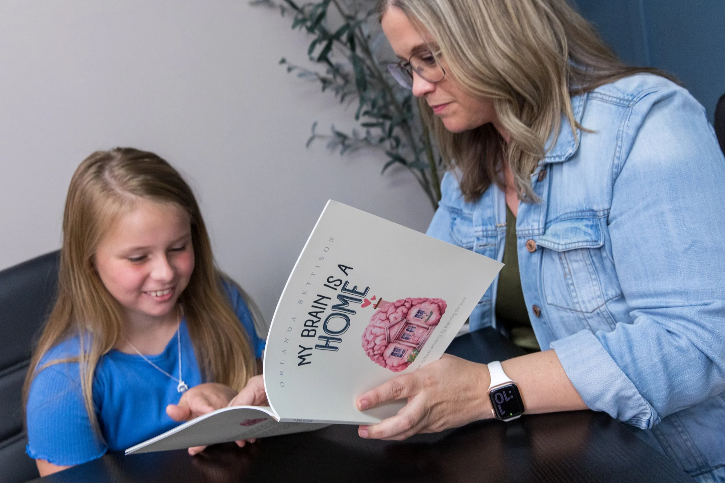 A woman and a young girl looking at a book titled 'My Brain is a HOME' with a pink house shaped like a brain on the cover, sitting at a table in a room with a plant in the background.
