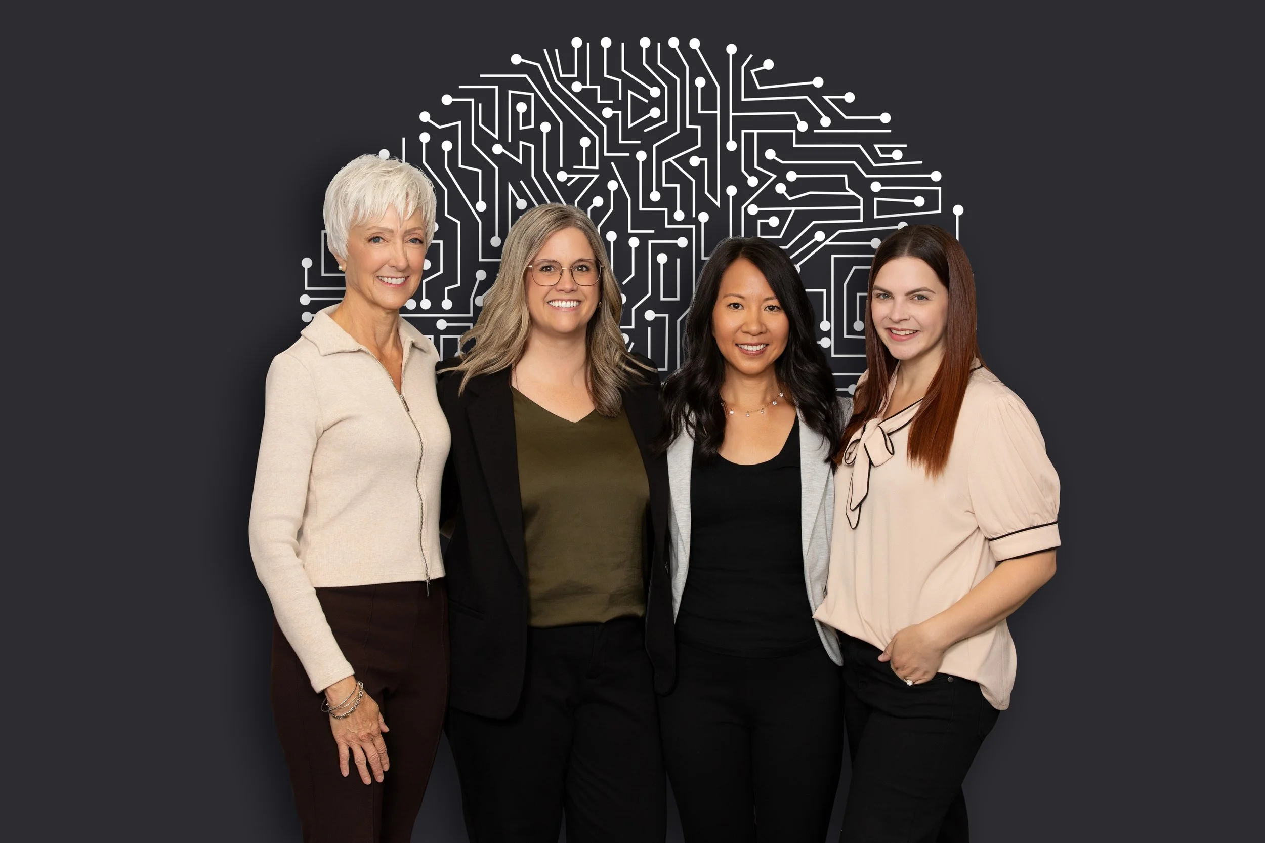 Four women standing together in front of a black background with a white circuit board design forming a brain shape. They are smiling and dressed in business casual attire.