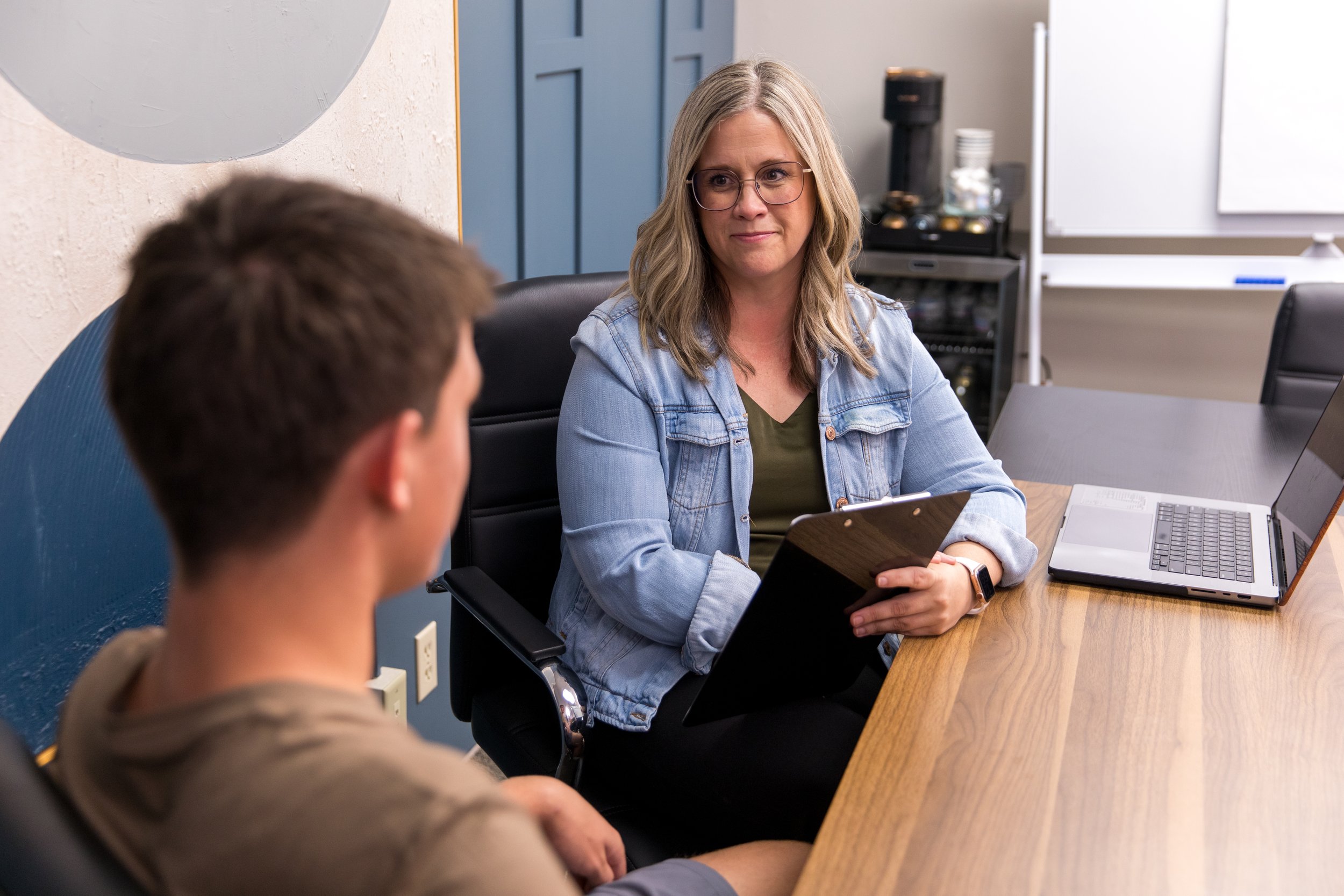 A woman with blonde hair and glasses wearing a denim jacket holding a clipboard, sitting at a desk, talking to a young man with brown hair, in a modern office setting with a laptop and coffee machine.
