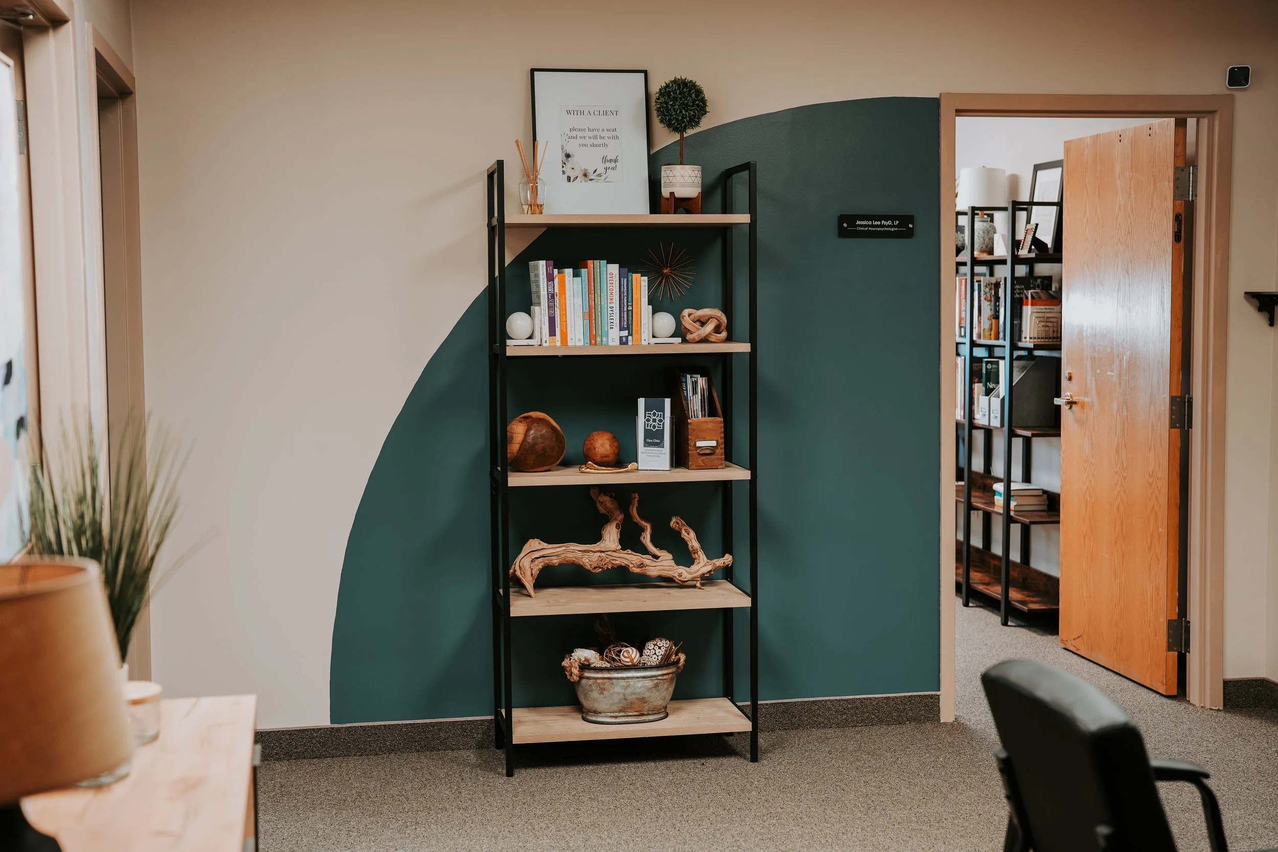 A black metal bookshelf with wooden shelves against a half green, half beige wall, displaying books, decorative objects, and a framed sign, with an open door to another room with bookshelves visible.