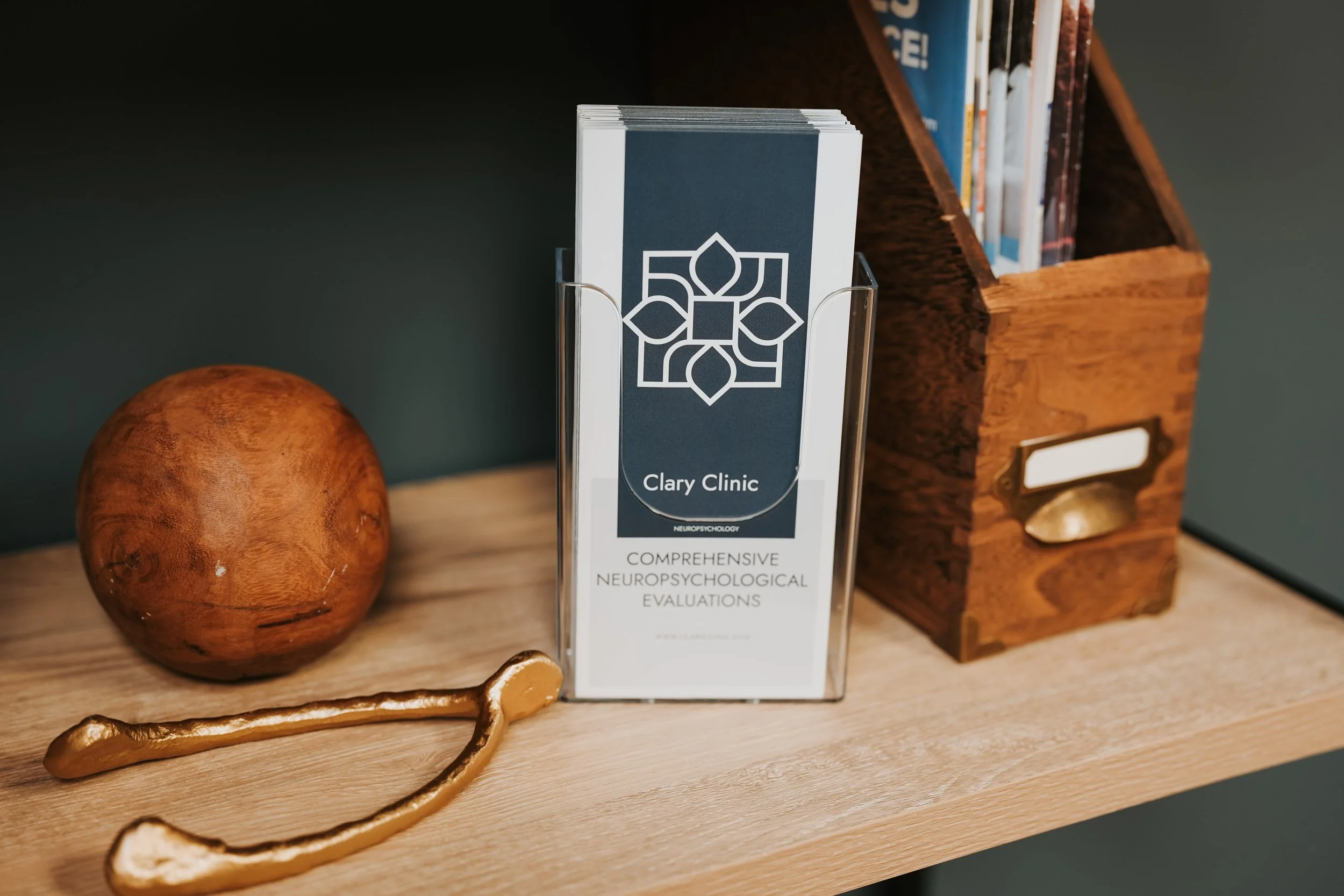Brochure for Clary Clinic with neuropsychology evaluations, a wooden ball, a gold-colored boomerang-shaped object, and a wooden file organizer with magazines on a wooden shelf.