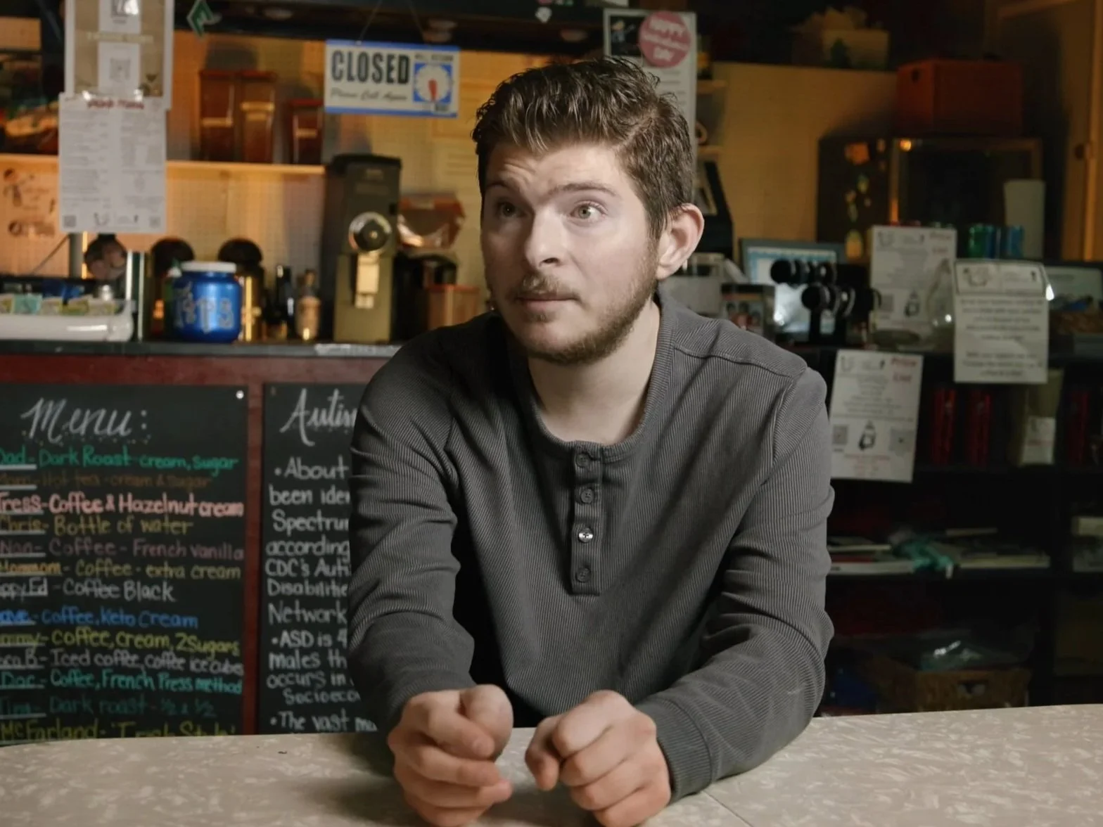 A young man with short brown hair and a beard sits at a counter inside a cafe, looking to his left. The cafe has a menu board with colorful writing behind him and various signs and items on the shelves.