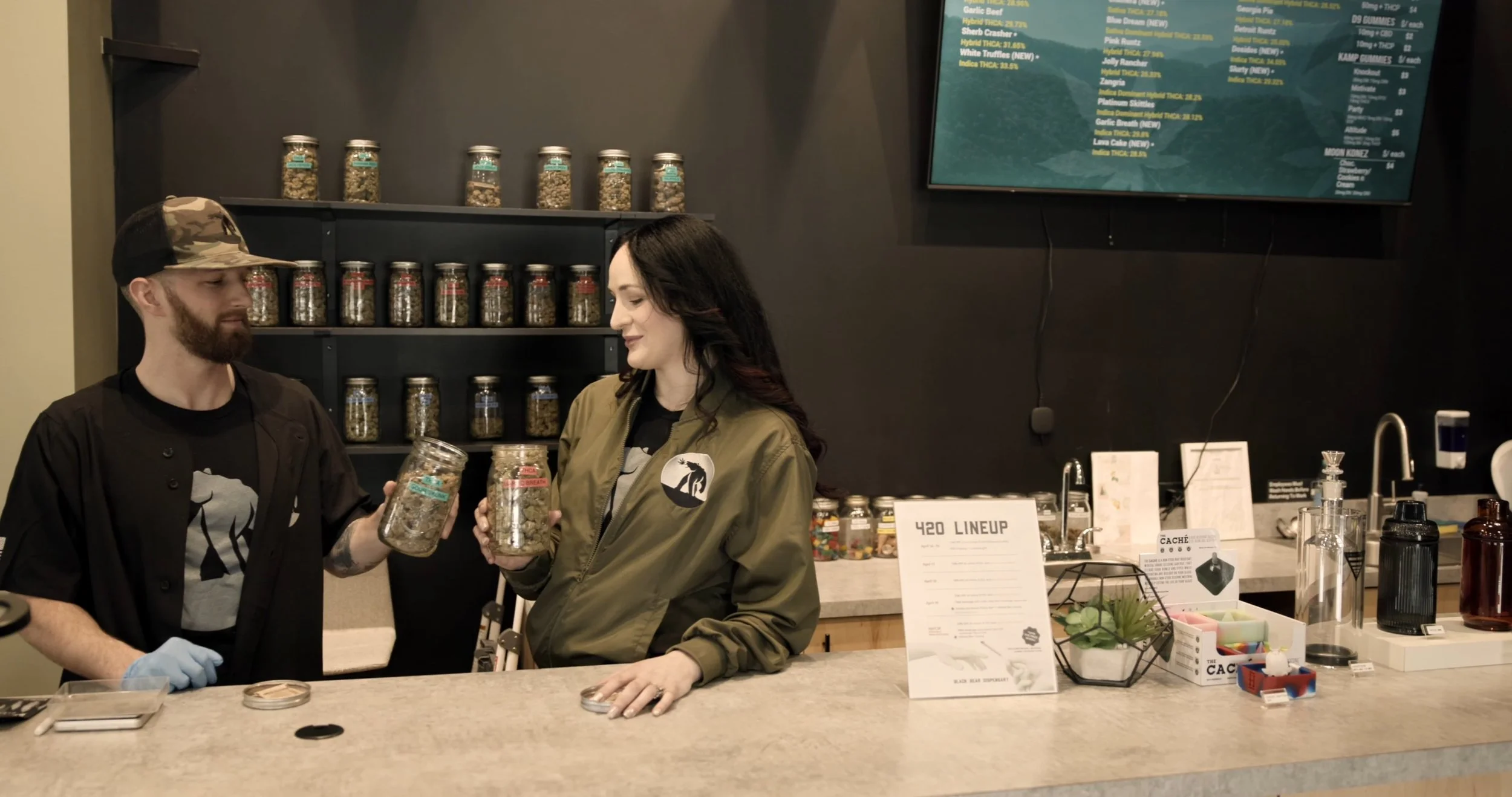 A man and woman are standing at a counter in a store, holding glass jars. The woman is wearing a green jacket with a black shirt underneath, and the man is wearing a black shirt, a camouflage cap, and has a beard. They are exchanging jars, and behind them are shelves filled with similar jars and a large digital menu on a wall.