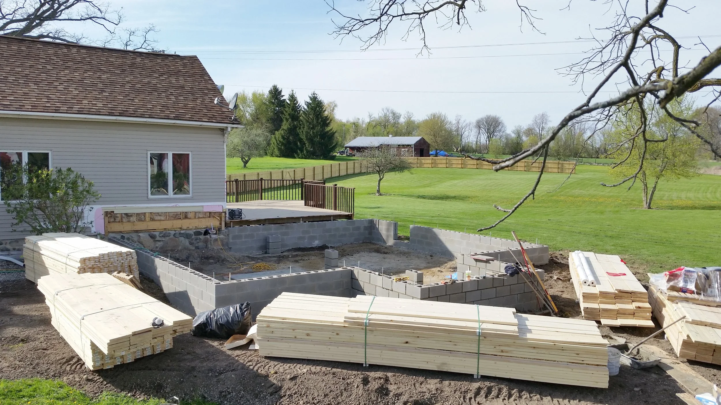 Construction site outlining a backyard with building materials, concrete blocks, and wooden panels on the ground, with a house on the left, and a grassy yard with trees and a barn in the background.
