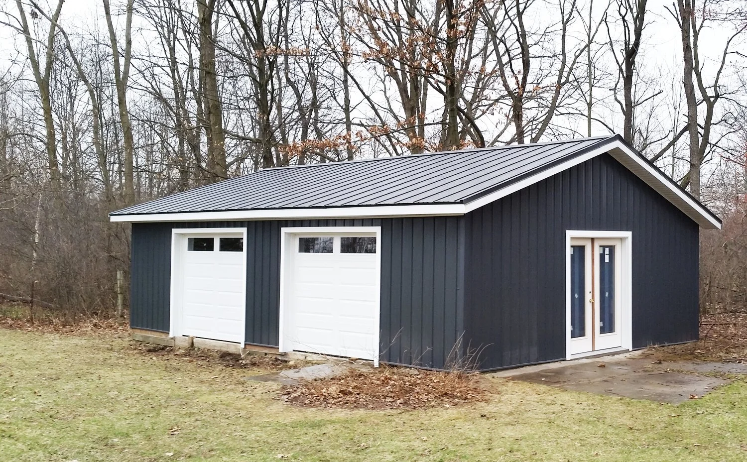 A black metal garage with two white garage doors and a white-framed glass double door, situated on a grassy yard with a wooded background.