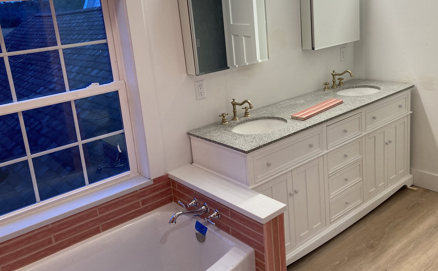 Bathroom with a double sink vanity with gold fixtures, white cabinets, and a granite countertop, beside a bathtub with a large window showing a tiled roof outside.
