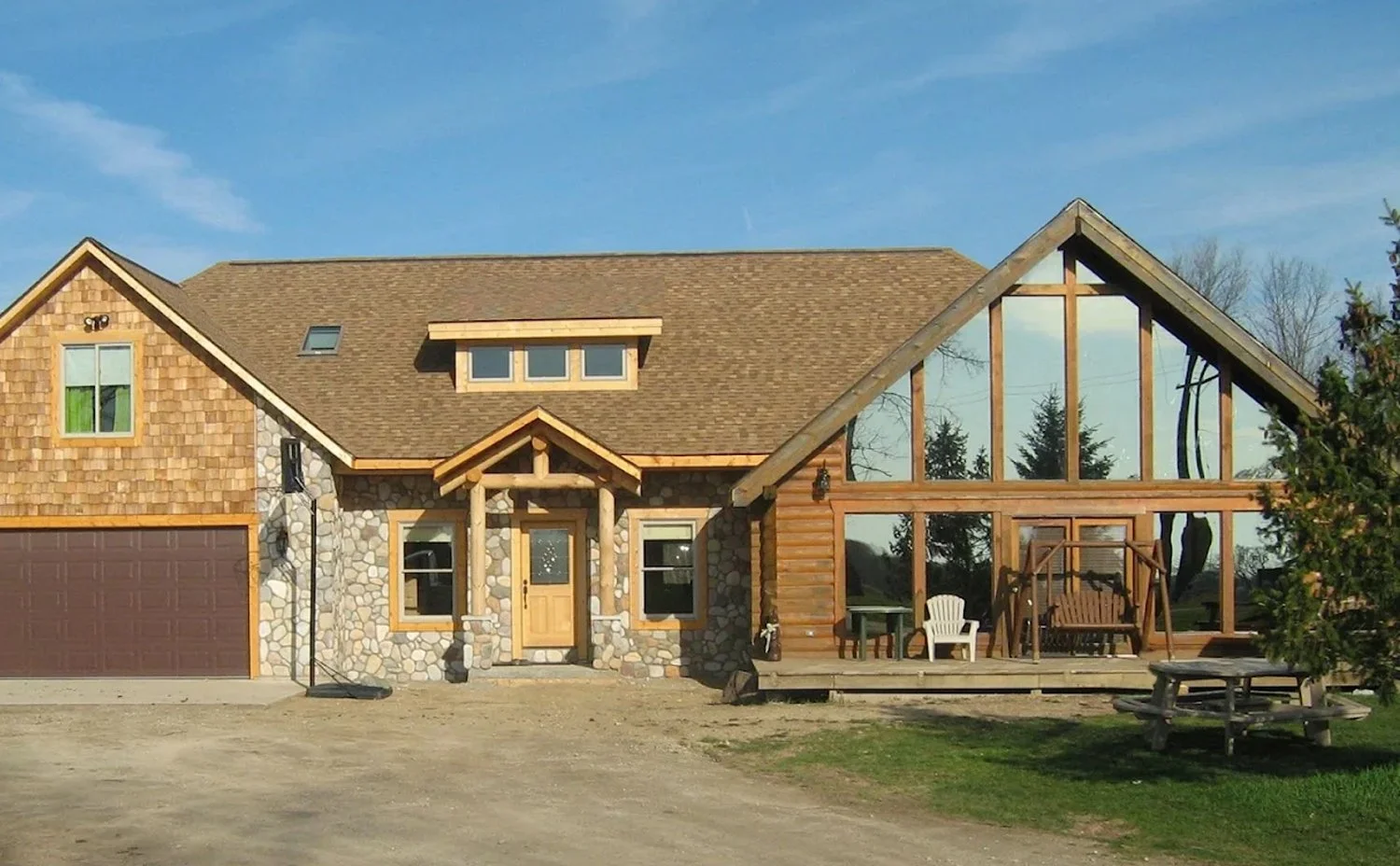 A house under construction with a large front porch. The house has a stone and wood exterior, with a brown roof and some Windows. The right side of the house shows a framed addition with large windows and outdoor furniture.