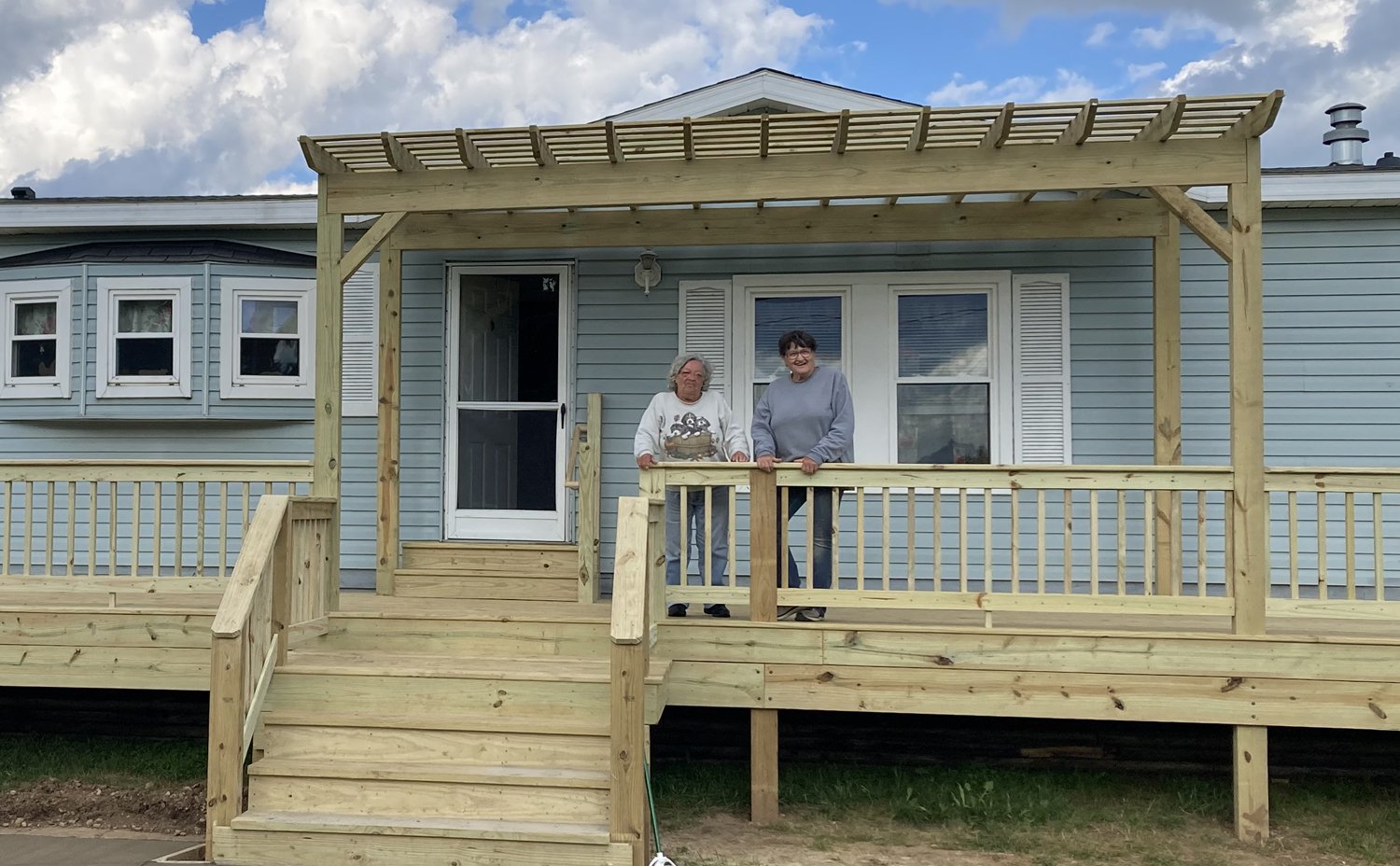 Two women stand on a newly built wooden deck in front of a light blue house, smiling at the camera. The deck has railings and a pergola on top.