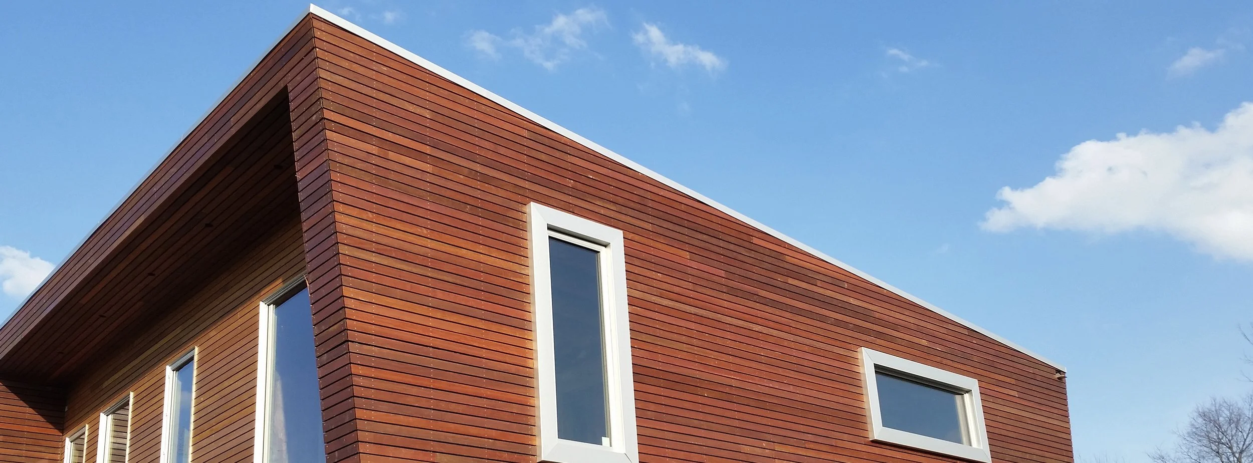 Modern building with reddish-brown wooden siding and white framed windows, against a blue sky with scattered clouds.