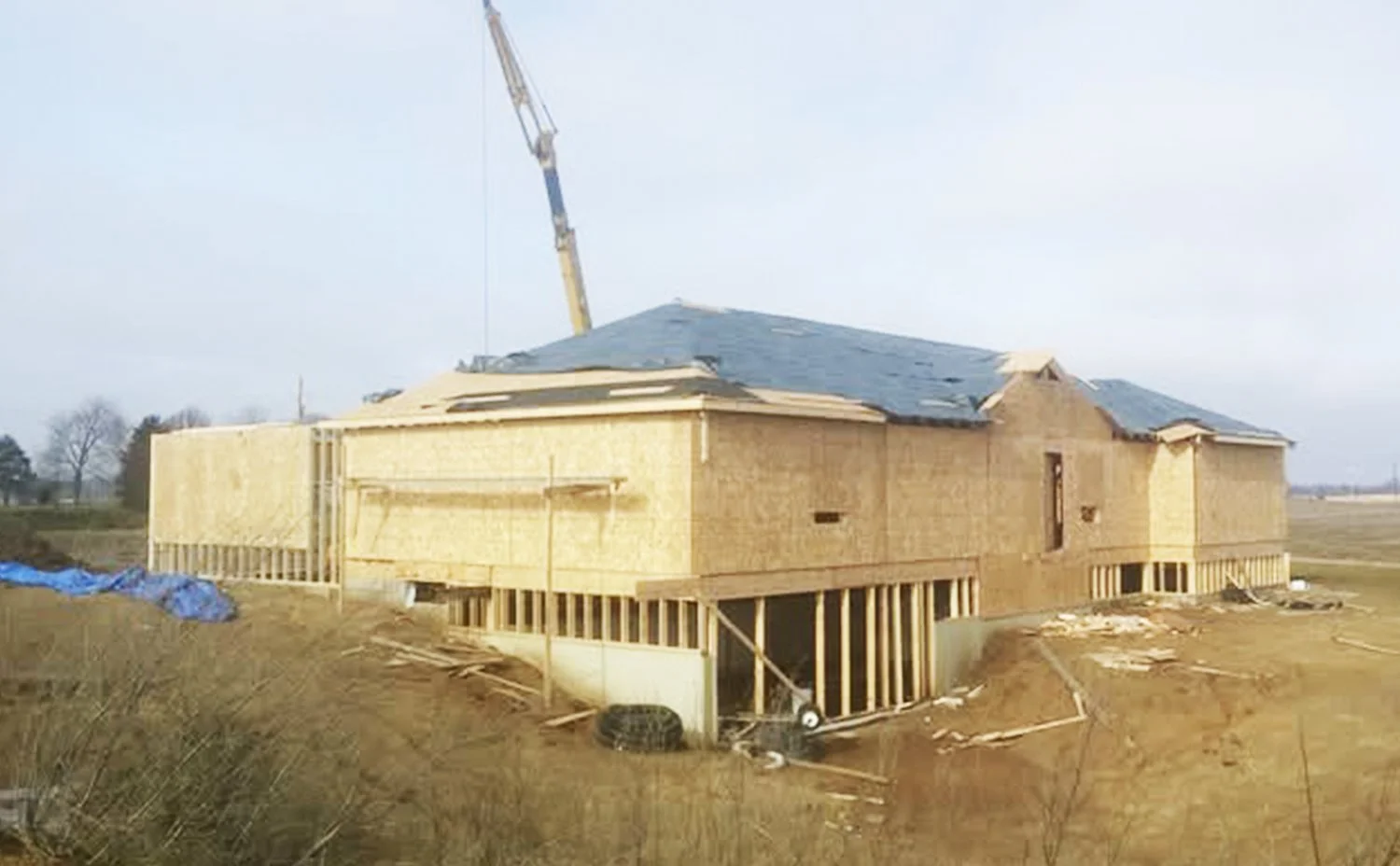 Under construction house with wooden framing and a blue tarp on roof, in an open field.
