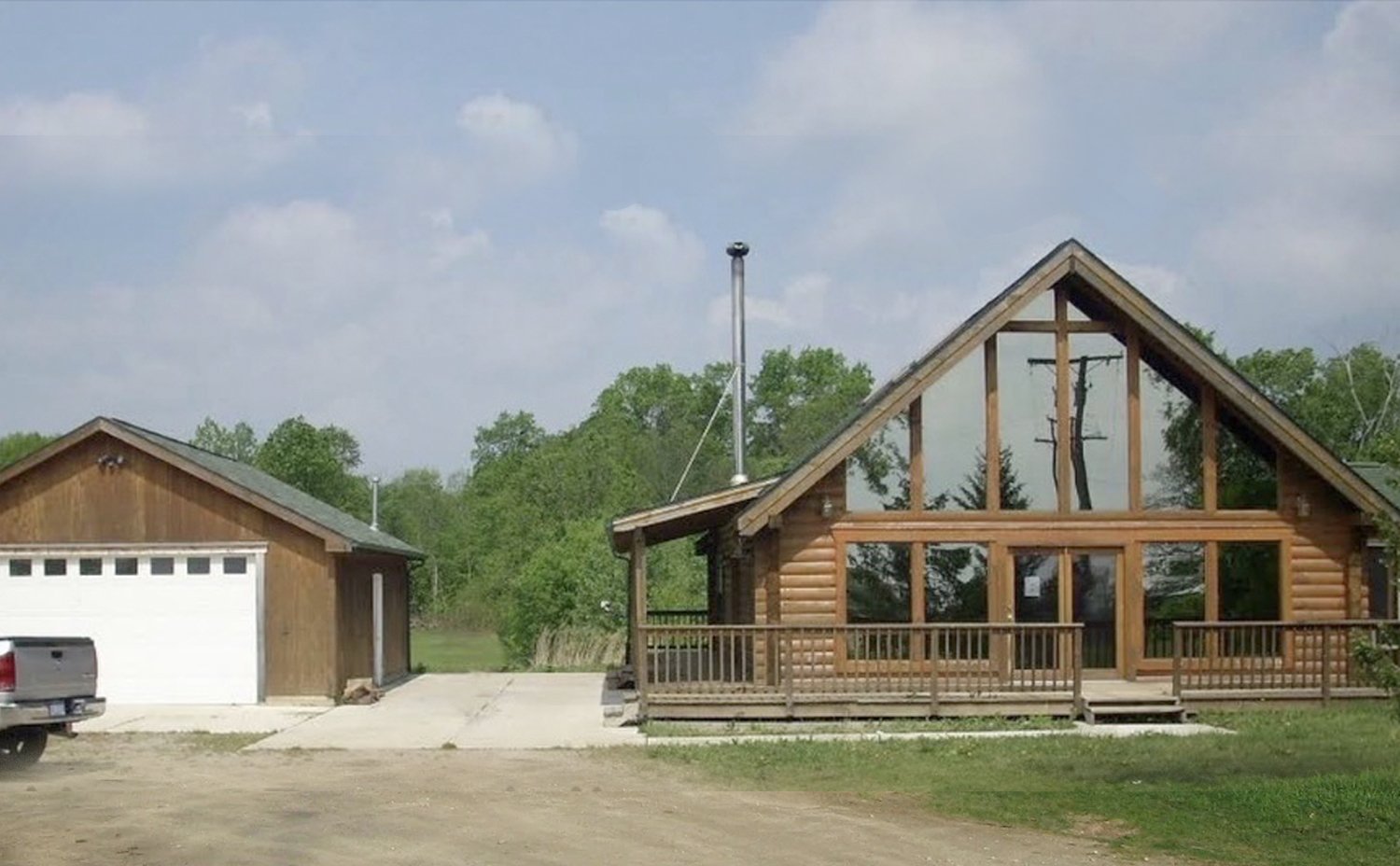 Log cabin with large front windows and a wooden balcony, situated next to a detached garage, with green trees and cloudy sky in the background.