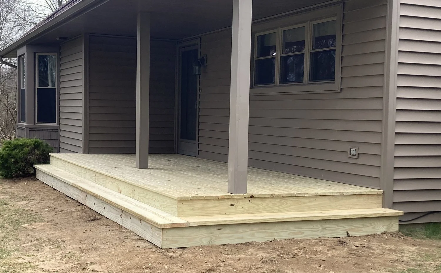 Newly built wooden deck attached to the back of a house with gray siding, featuring steps leading down to a dirt yard, supported by two vertical posts, with a windows and door leading into the house.