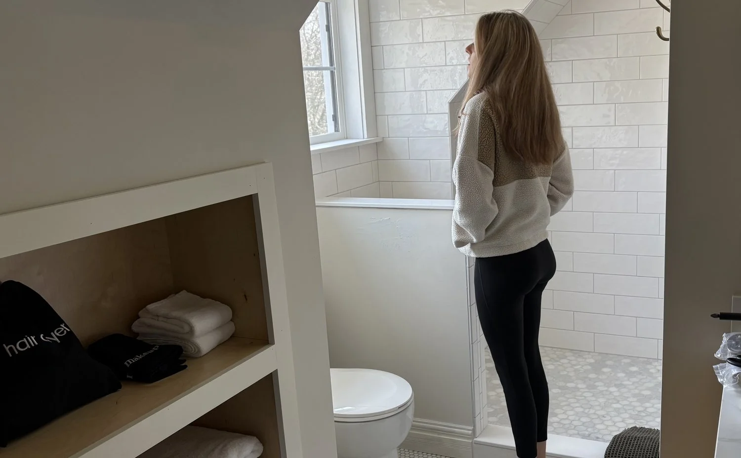 A woman with long hair looking out a window in a modern bathroom with white tile walls and a pebble tile floor.