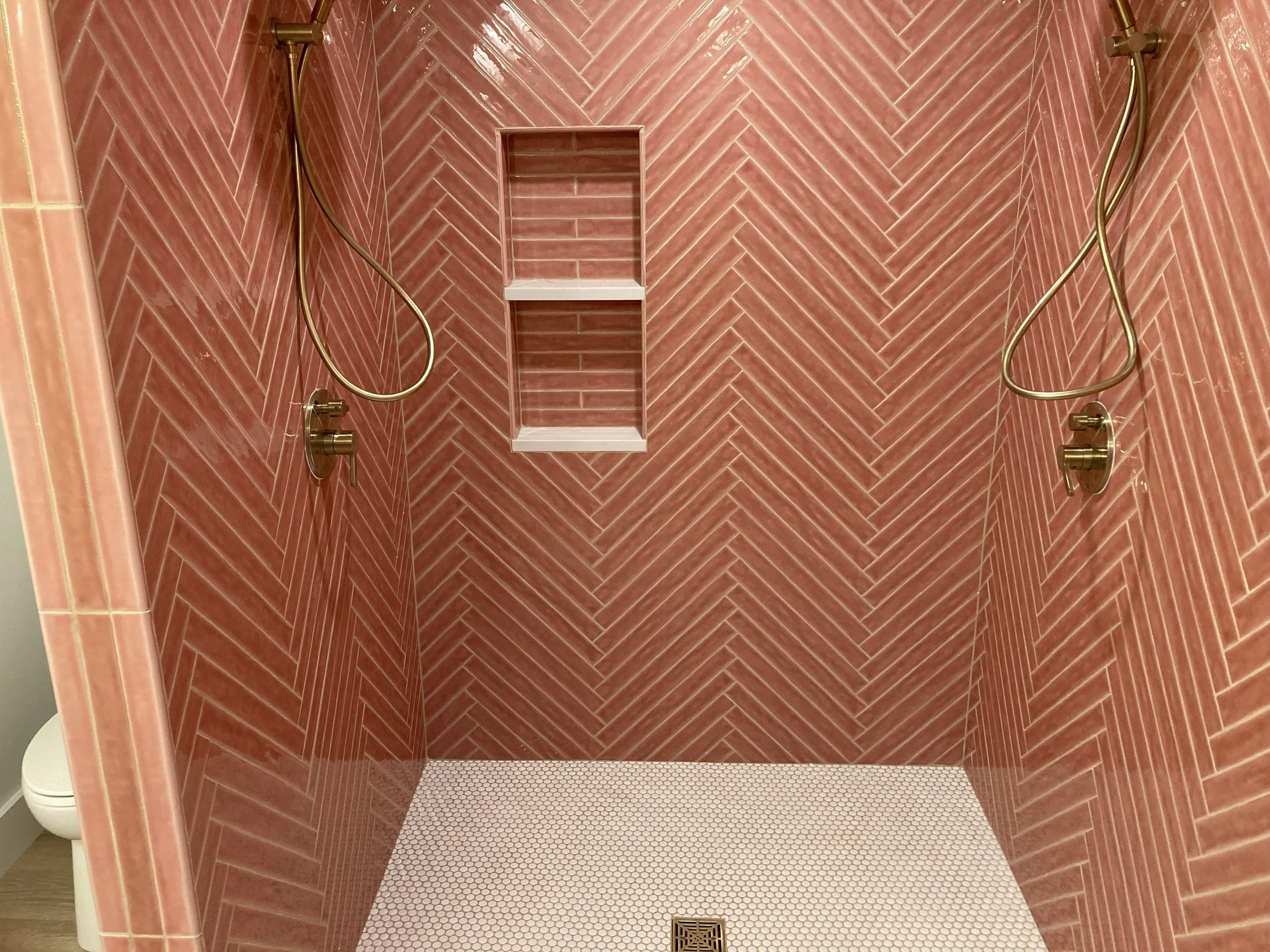 A shower area with pink tile walls arranged in a herringbone pattern, two brass showerheads, a small built-in shelf with three tiers, and a white tile floor with a central drain.