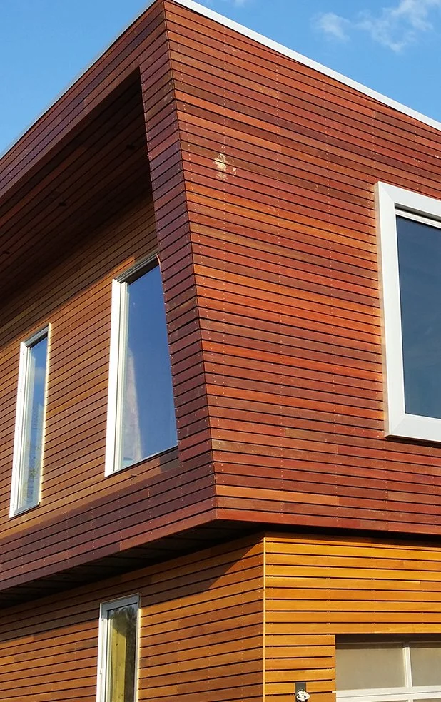 Close-up of a modern building's corner with brown and orange wooden siding and white-framed windows, with a blue sky in the background.