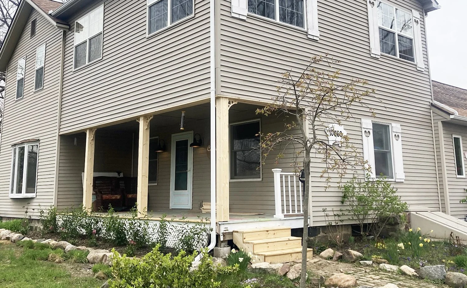 A two-story house with beige siding and white shutters; porch with wooden stairs, a small tree in front, and garden plants around.