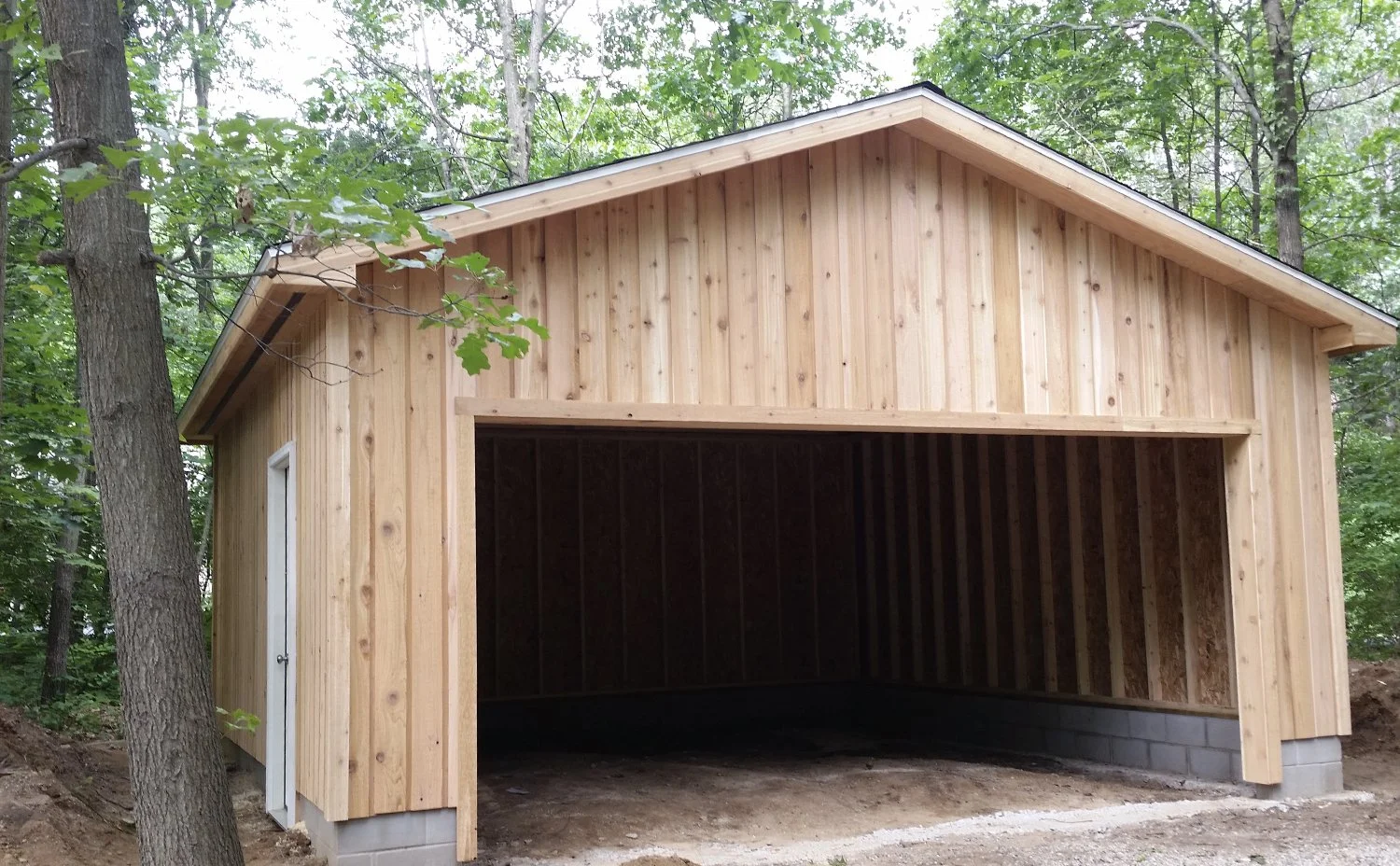 A newly built wooden garage in a wooded area, with an open front and a small side door, showing exposed interior framing.