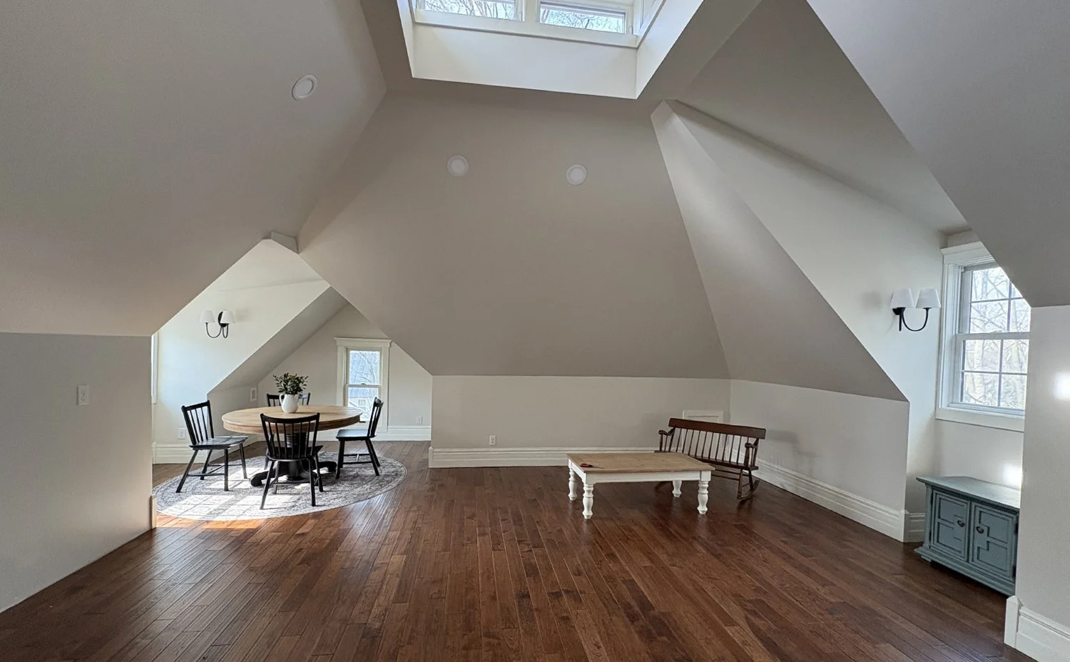 Spacious attic room with sloped ceilings, hardwood floors, two windows, a small round dining table with chairs, a coffee table, a bench, a cabinet, and wall-mounted light fixtures.