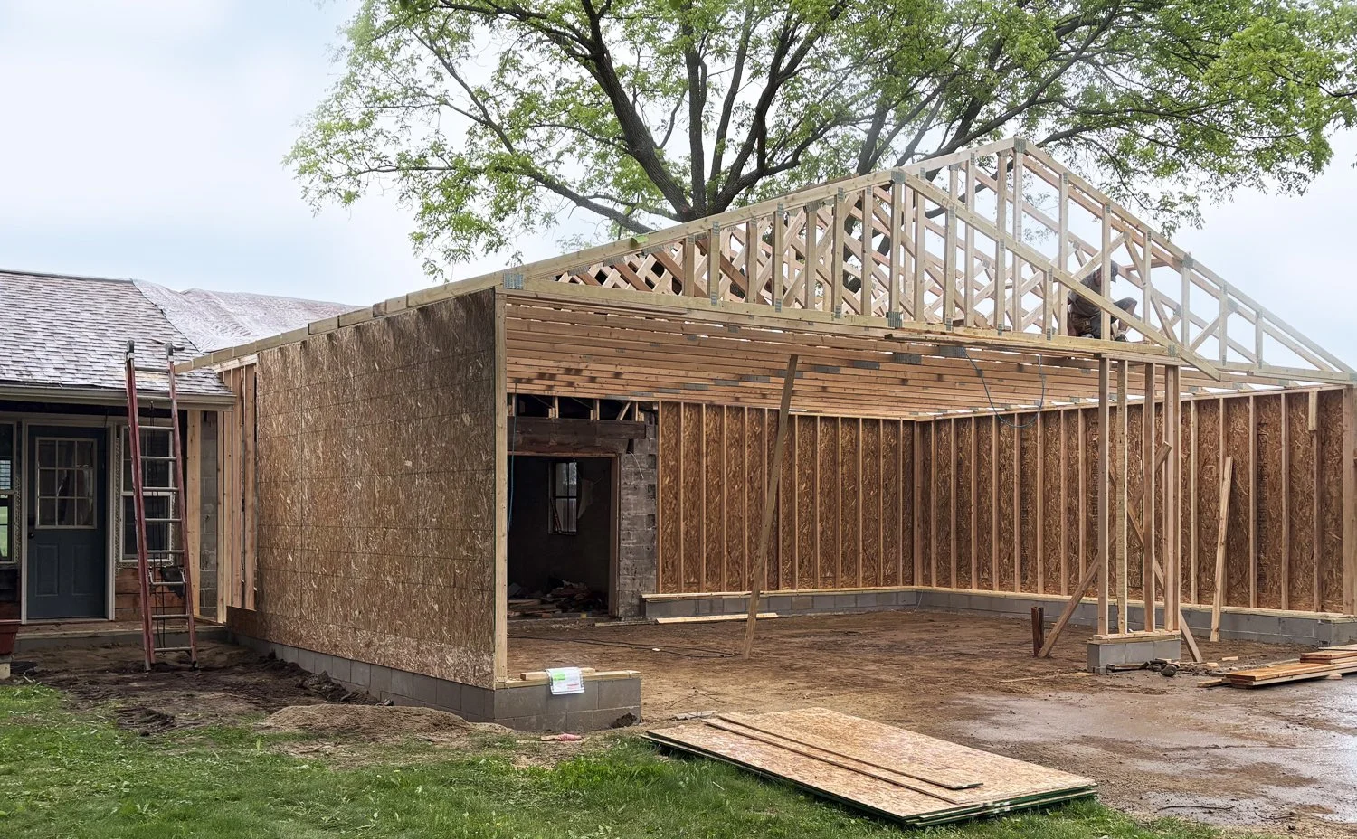 Construction site of a new building with wooden framing and walls in progress, next to an existing house with a louvered door, a ladder leaning against the house, and trees in the background.