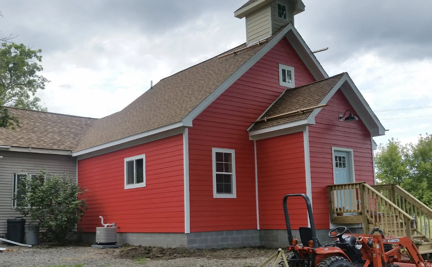 A red barn-style house with white trim, a small blue door, and a small set of wooden stairs leading to the entrance. There is a Kubota tractor parked in front of the house, and some trees and a cloudy sky are in the background.