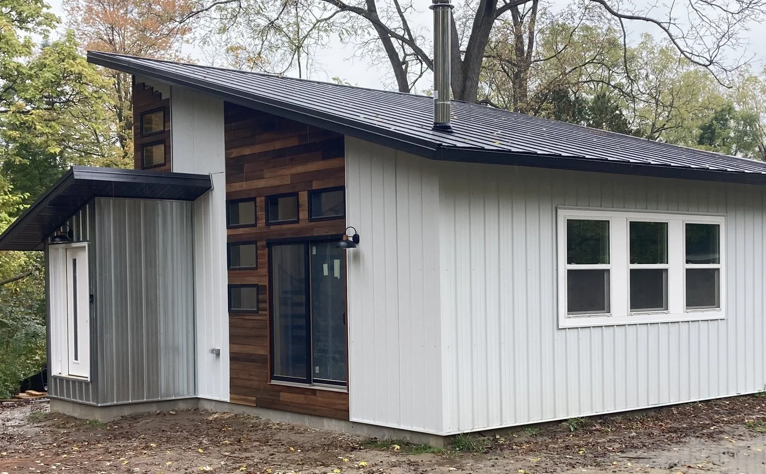 A modern house with white metal siding, dark metal roof, and a wood-paneled section with a large sliding glass door. There are four small windows above the door, a black outdoor light, and a corner window on the side.