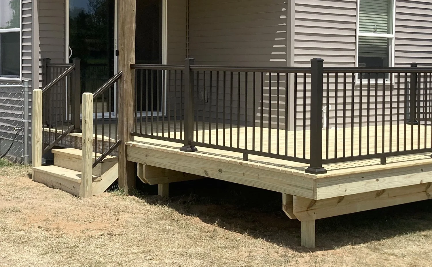 Newly built wooden deck with black metal railing attached to a house with beige siding, and small wooden stairs leading down to dry grass.