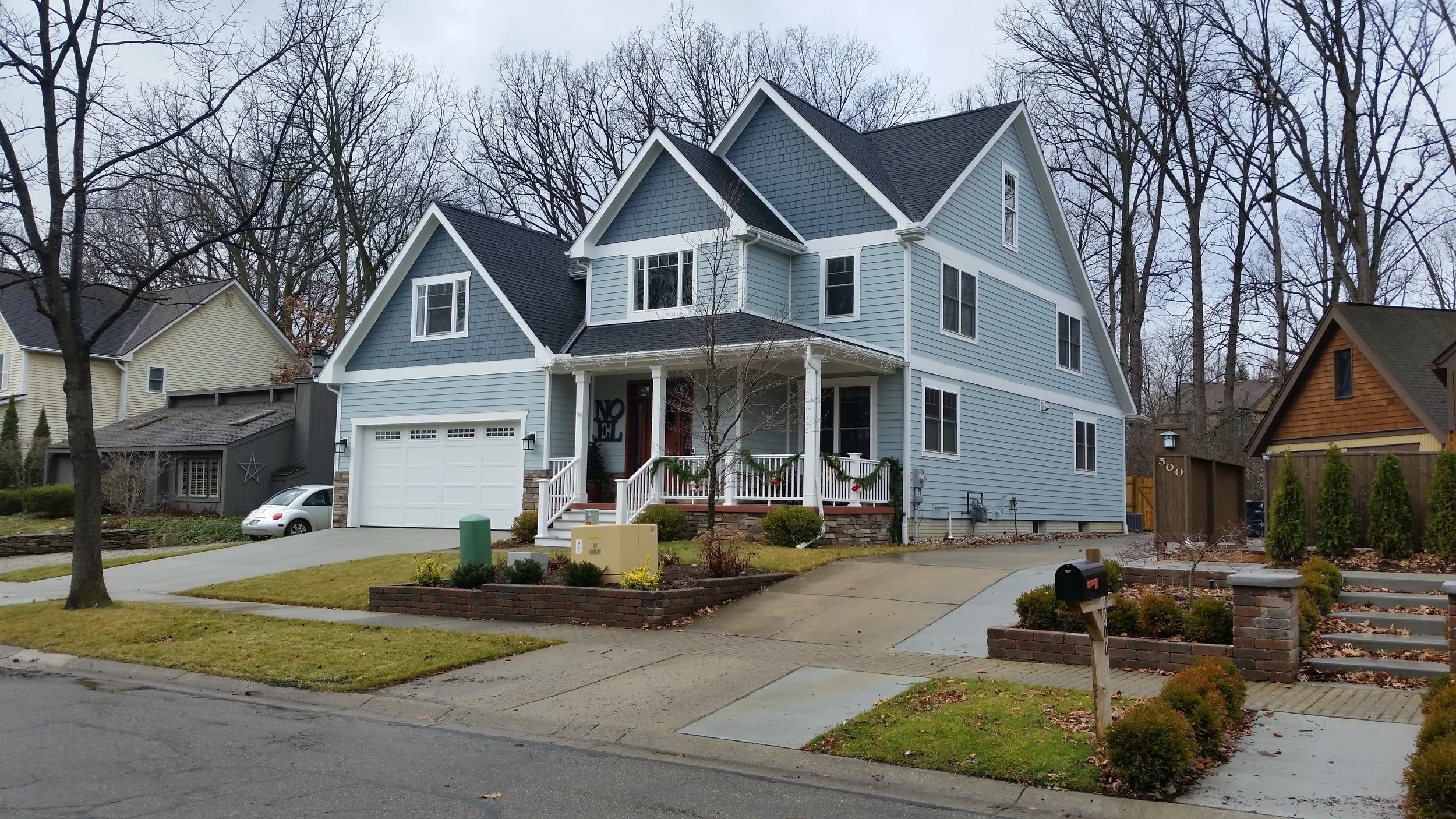 A large, two-story blue house with white trim and a front porch decorated for Christmas, located in a suburban neighborhood with neighboring houses and leafless trees in the background.