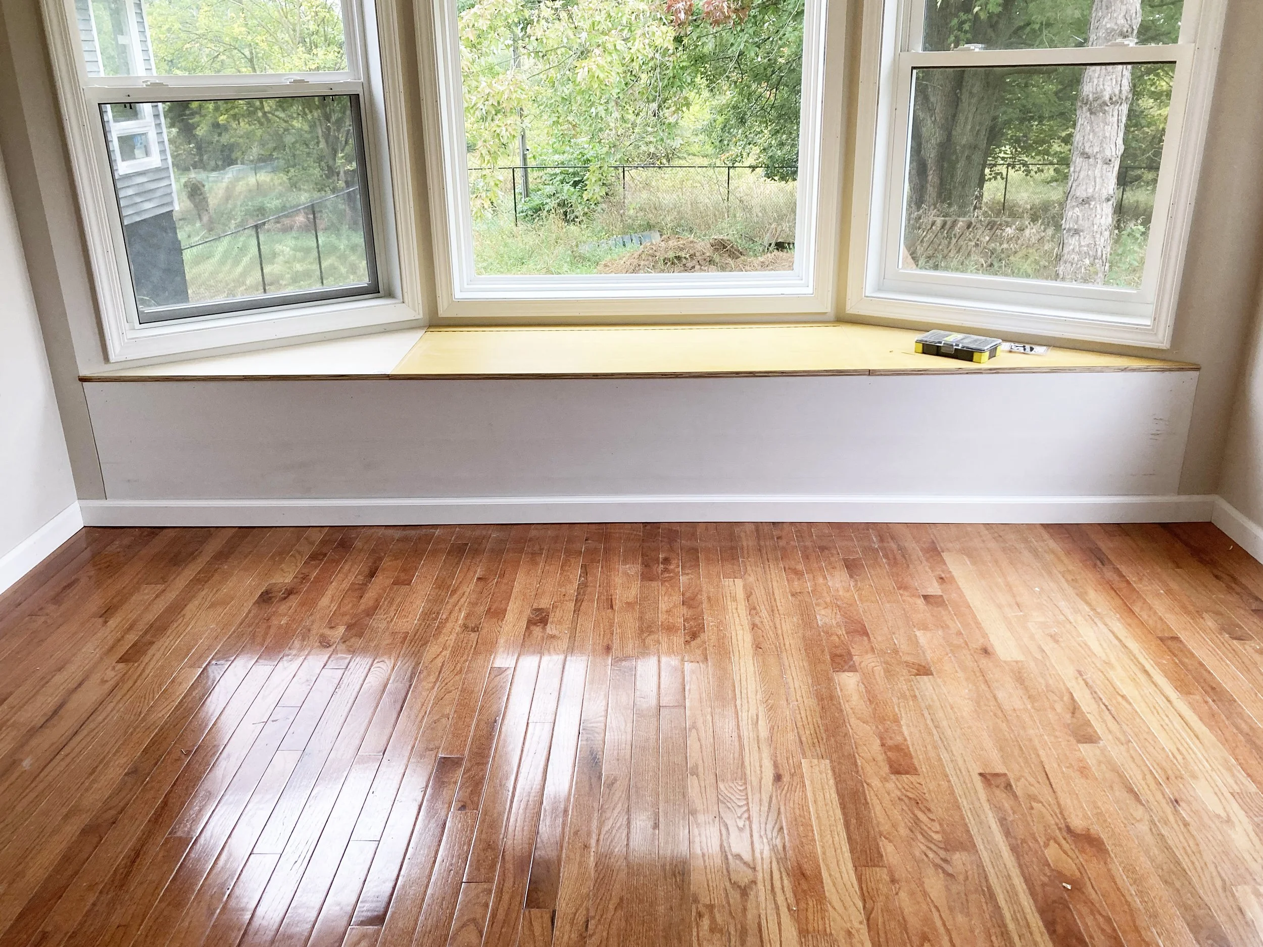 Empty room with hardwood floor, three large windows with white frames, and a window seat with yellowish top. There are tools and a tape measure on the window seat, and trees and greenery visible outside.