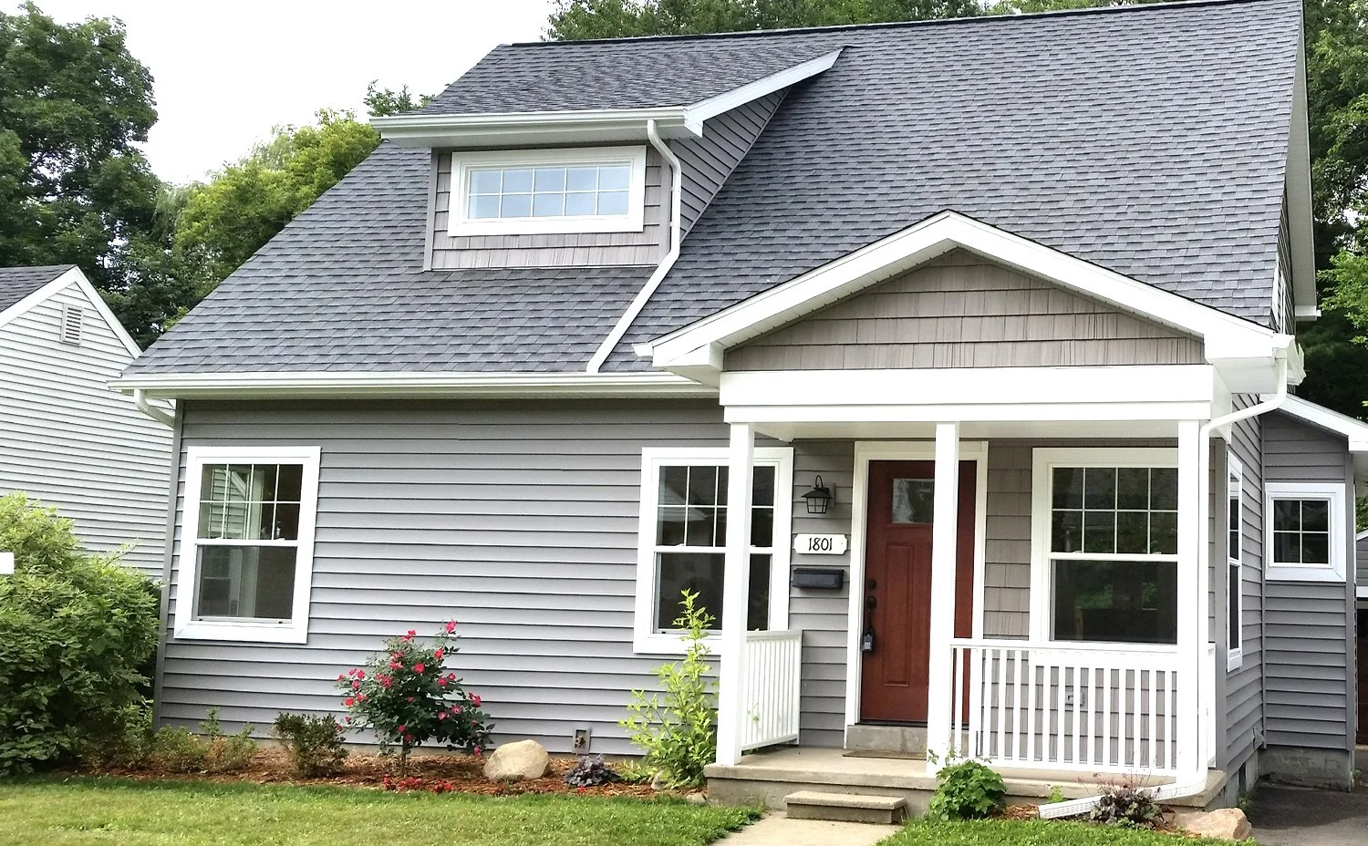 Gray two-story house with a small front porch, a red door, and multiple white-framed windows, surrounded by a landscaped lawn with shrubs and flowers.