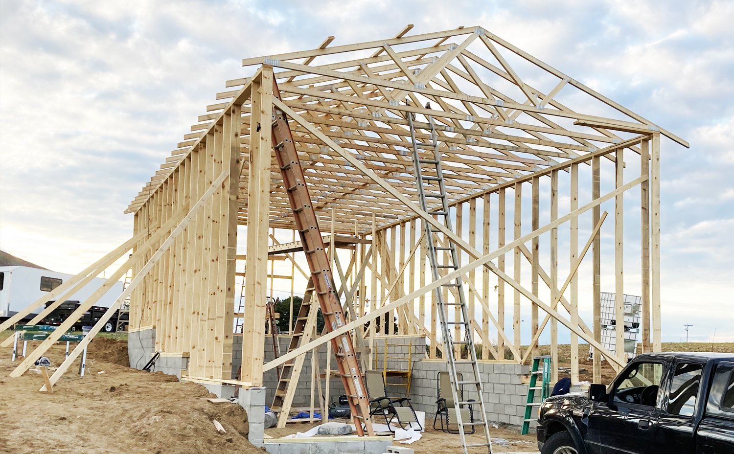 Wooden framing of a building under construction with ladders and construction equipment, set on a dirt lot with a partly cloudy sky.