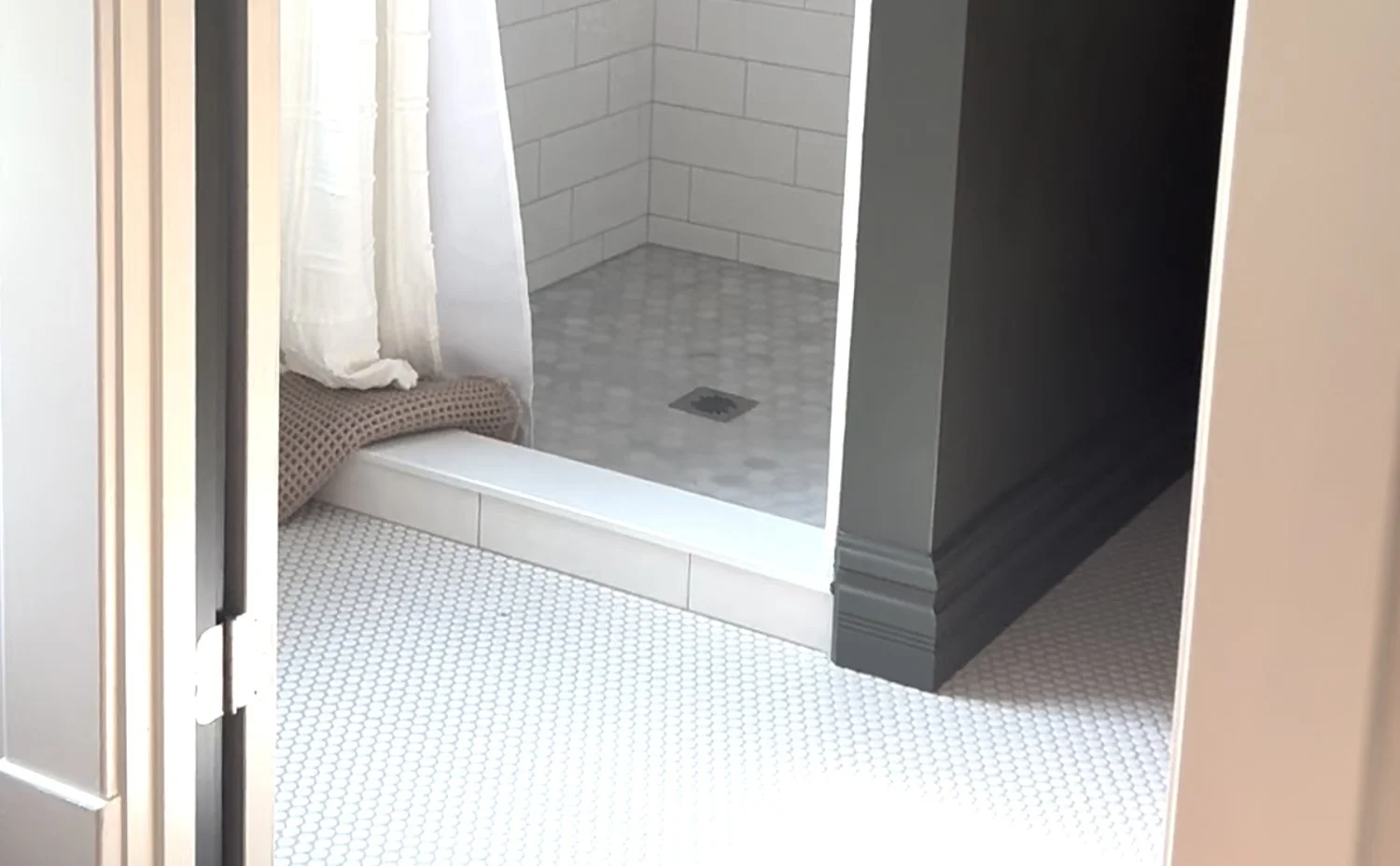 Partial view of a bathroom with a shower area with white tiled walls and a drain on the floor. There is a curtain and some towels or cloths on the edge, and part of a black cabinet or wall is visible.