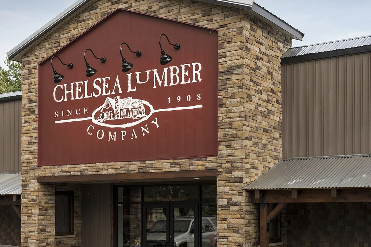 Exterior view of Chelsea Lumber Company building with a large red sign displaying the company's name, since 1908, and a graphic of a barn, with brick and metal siding walls and a glass door entrance.