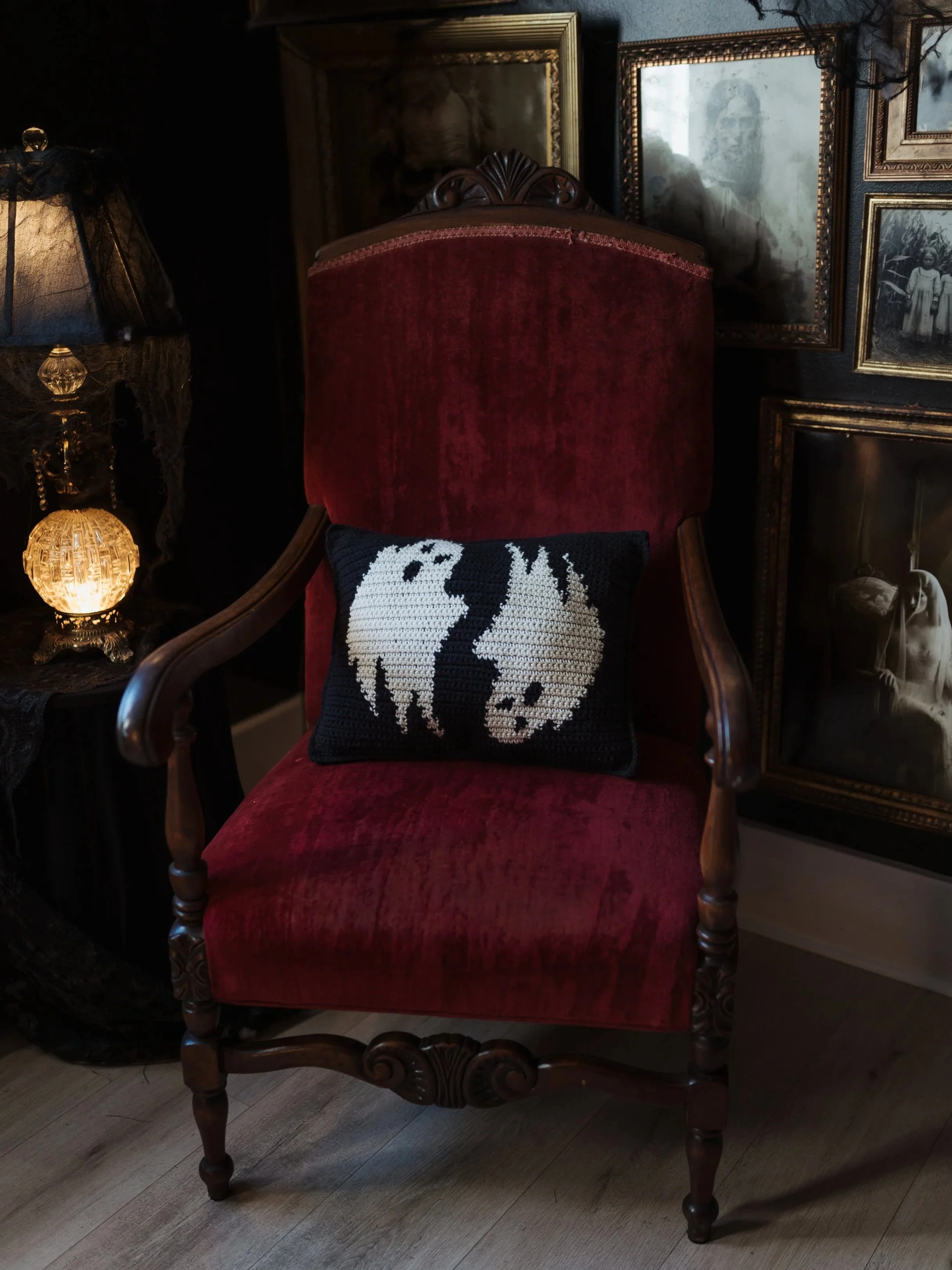 A vintage red velvet armchair with a decorative wooden carved frame, placed in a room with dark walls. Behind the chair, there are framed black-and-white photographs and artwork on the wall. A crochet pillow with spooky design rests on the chair.