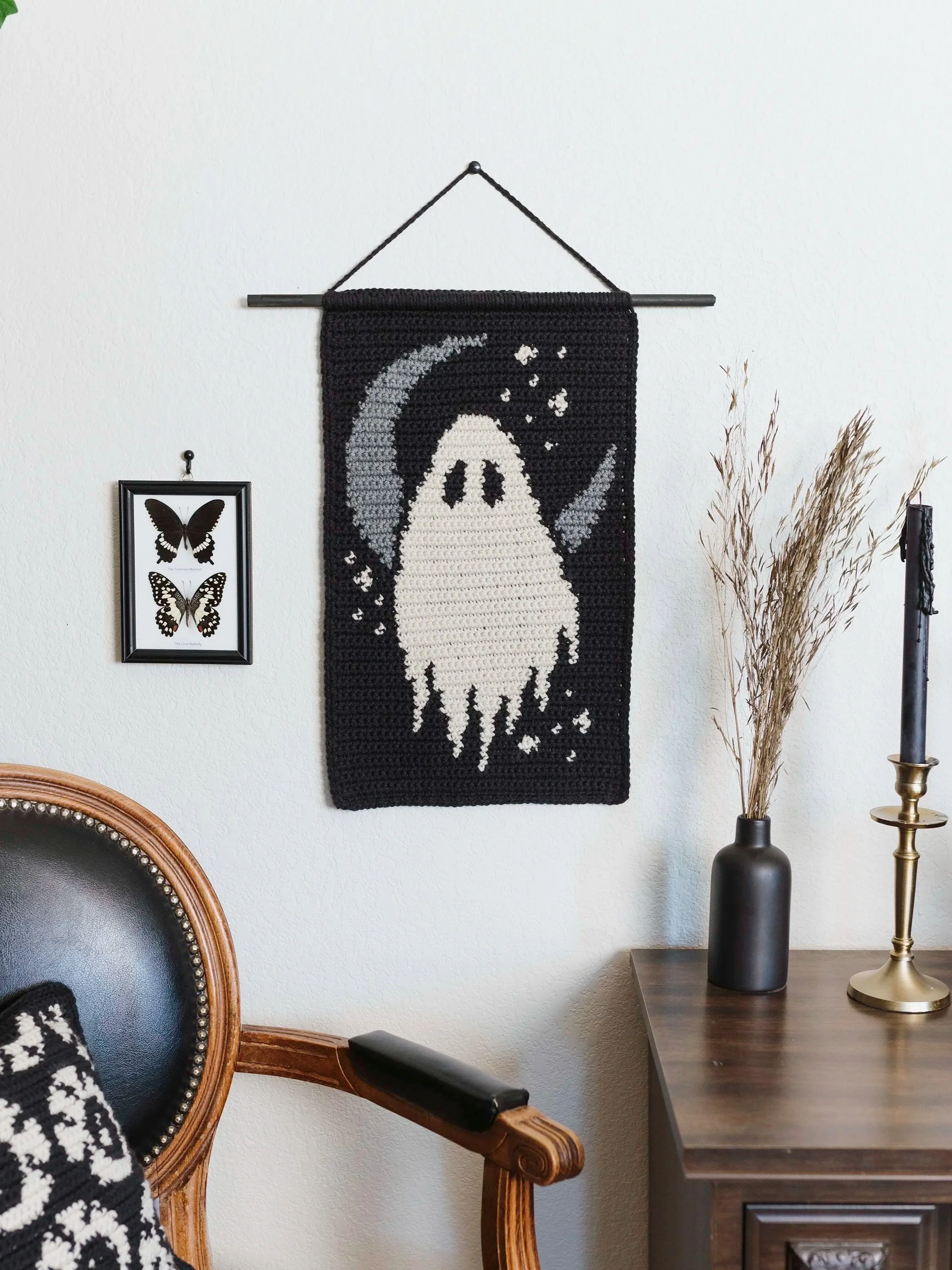 A black crocheted wall hanging with a ghost, moon, and stars design, hung on a decorative black rod with a string on a white wall, with a framed butterfly artwork to the left and a black vase with dried grasses and a brass candlestick on a wooden tab