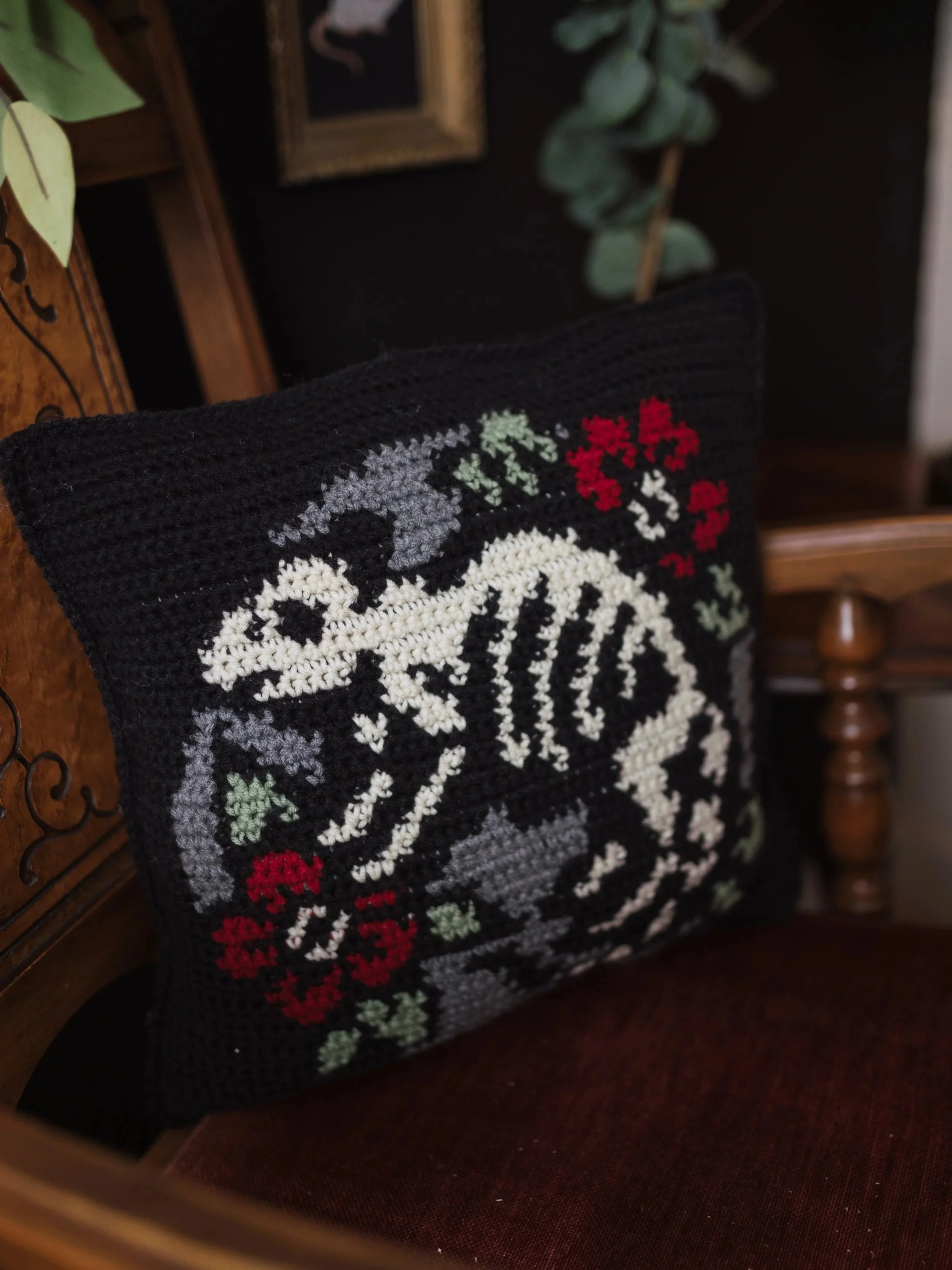 A black crochet cushion with embroidered white skulls, red flowers, and green leaves, resting on a dark red surface, with green leafy plants and a wooden frame in the background.