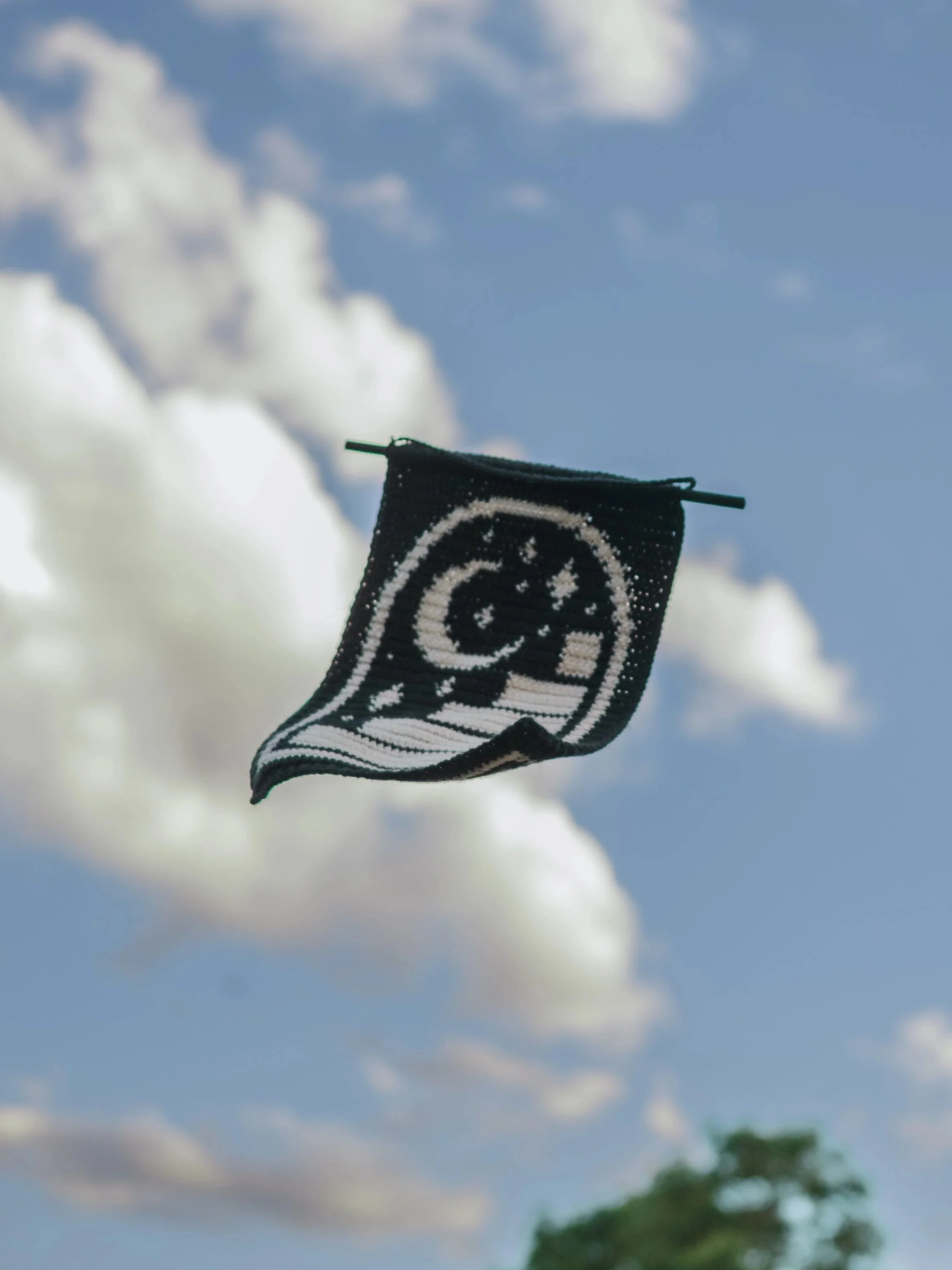 Black and white crochet wall hanging flying against a partly cloudy sky with cumulus clouds and trees in the distance.