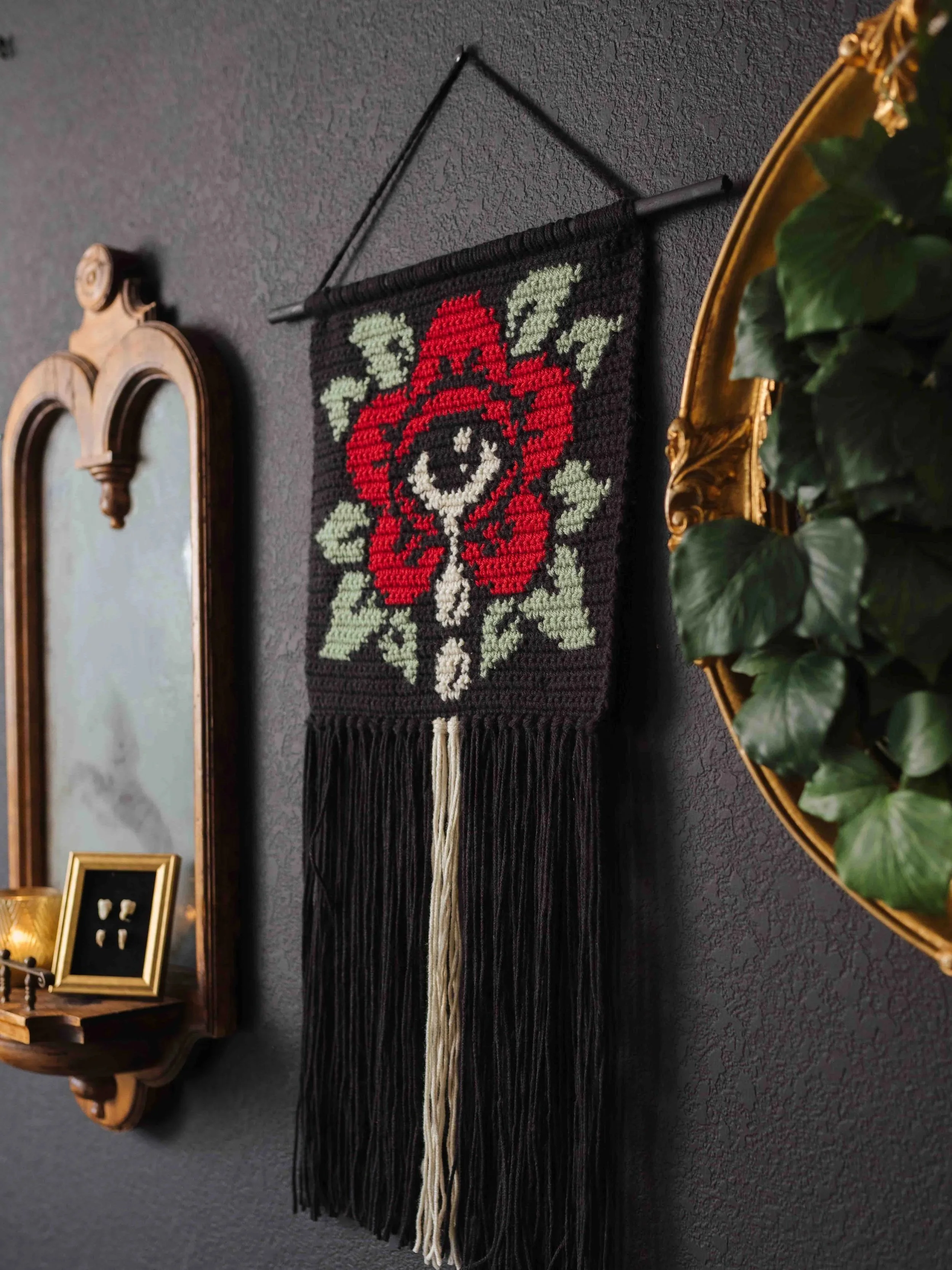 A black crochet wall hanging with a red and white floral pattern and green leaves, hanging on a dark gray textured wall, flanked by a gold-framed mirror and a small wooden shelf with decorative items.