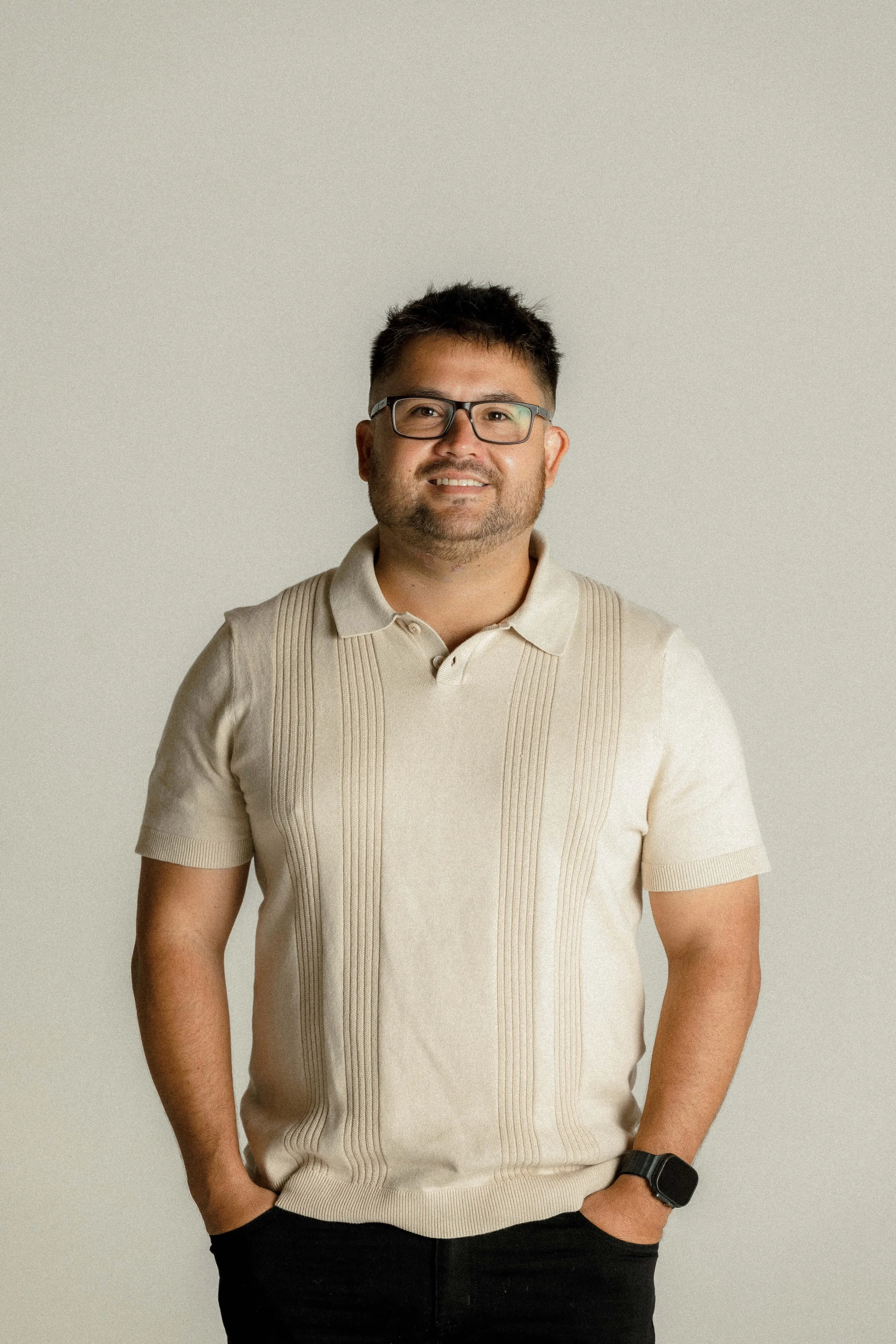 A man wearing glasses, a beige short-sleeve collared shirt with vertical stripe details, a black watch, and dark pants standing against a plain light gray background.