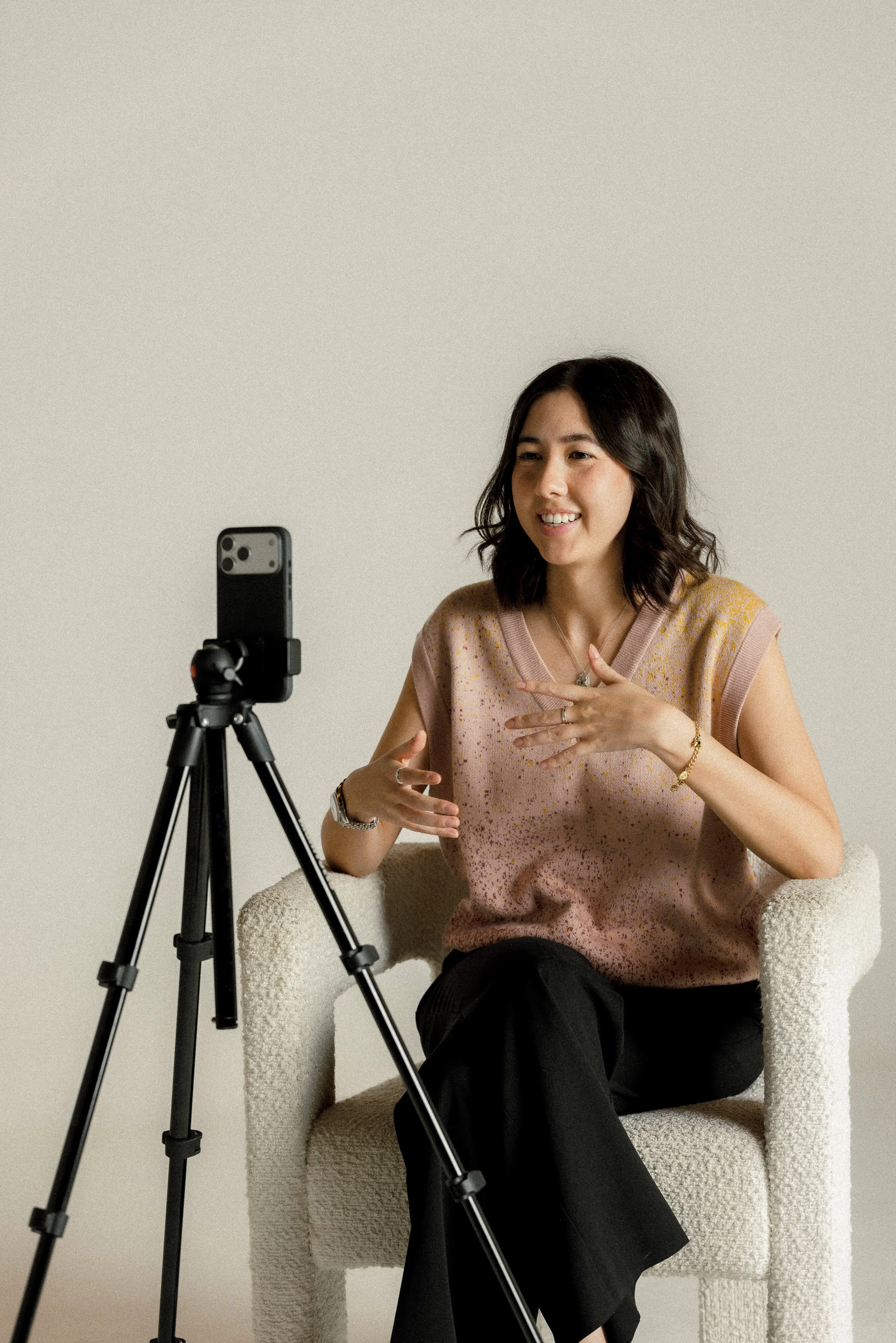 Kalya sitting on a white plush armchair, gesturing with her hands, talking to a phone mounted on a tripod for a video call or recording.