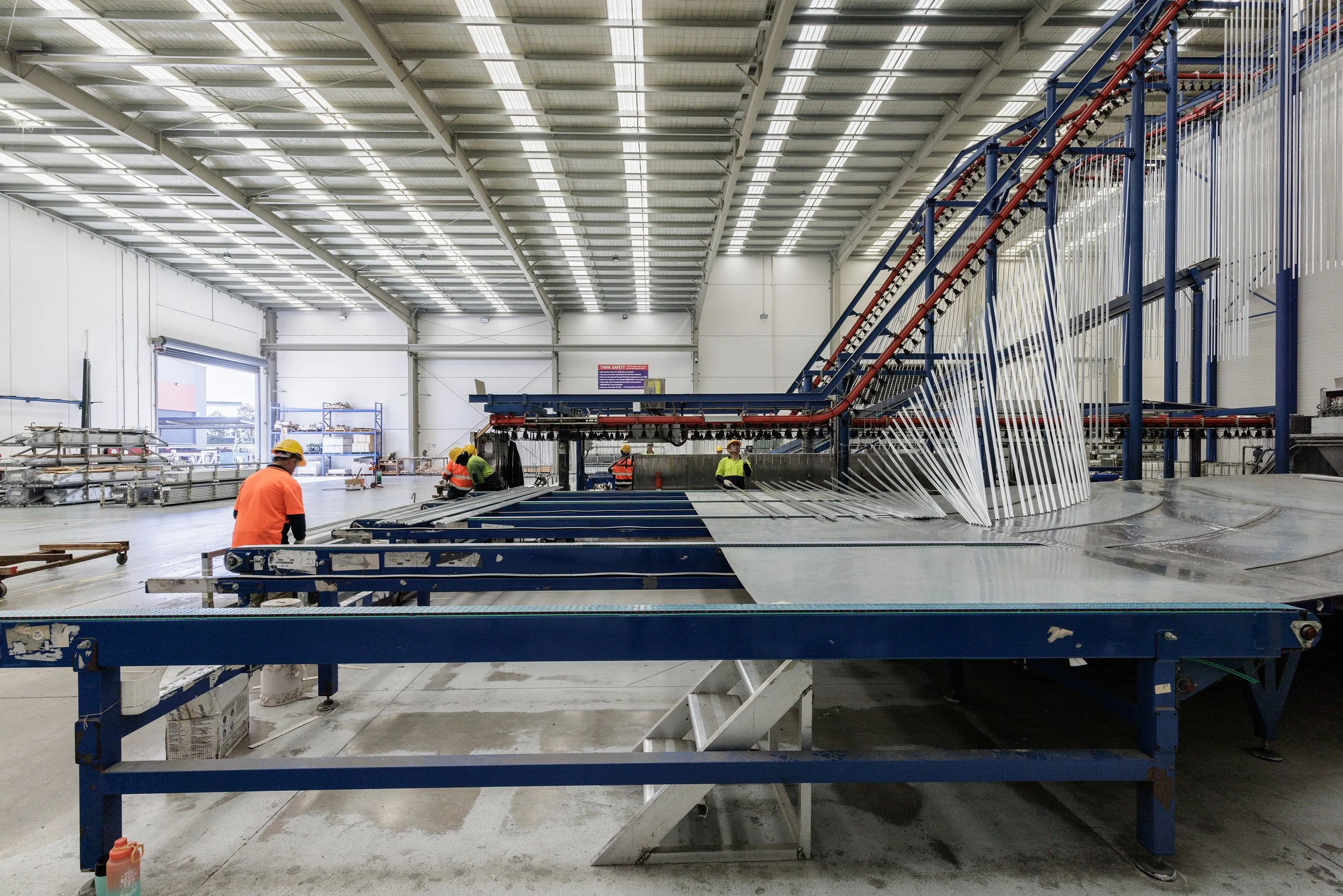 Inside a large industrial warehouse, workers in safety helmets and vests work near conveyor belts and machinery, with a high ceiling and bright overhead lighting.