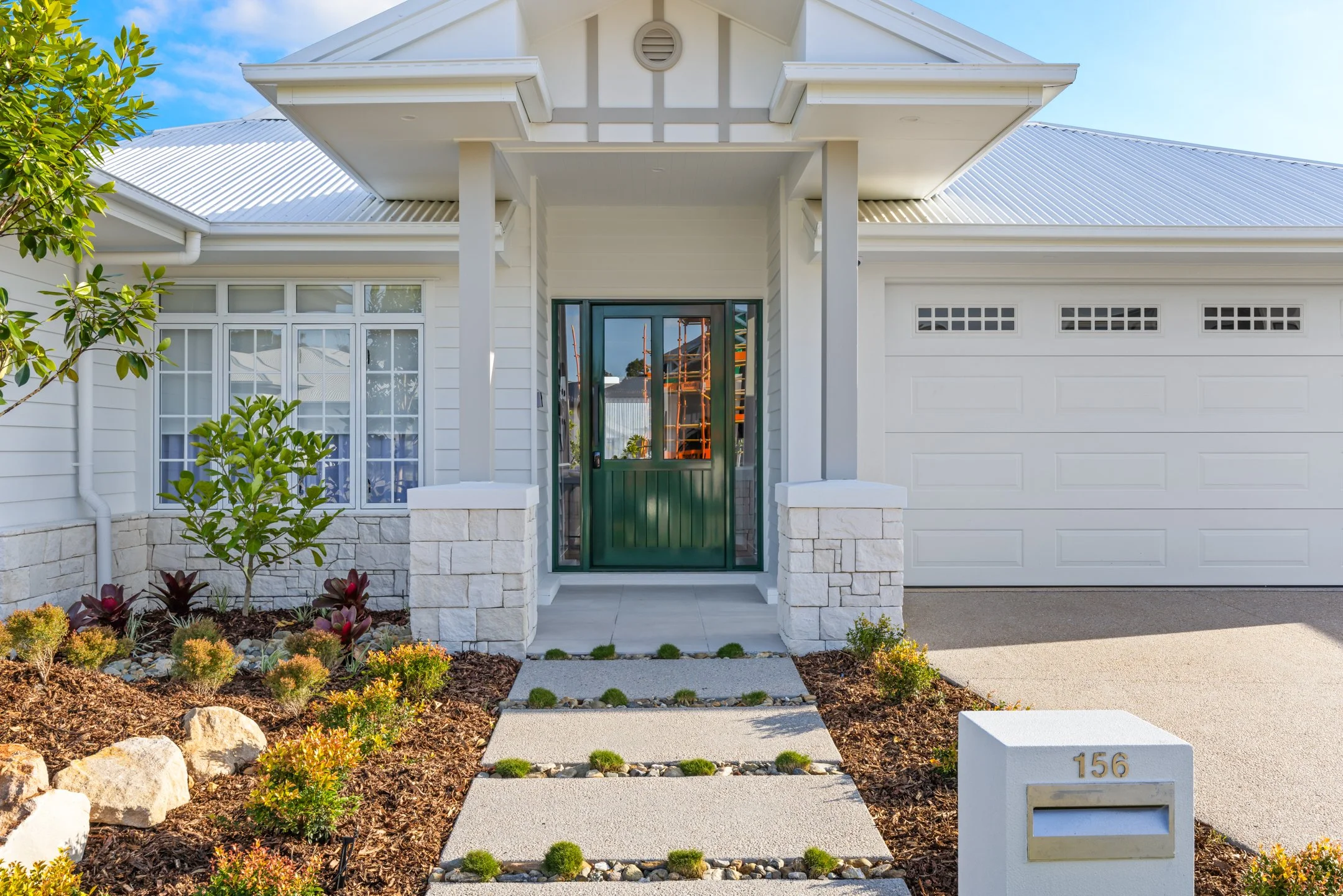 Front view of a modern white house with a grey roof, green front door with glass panels, landscaped garden with small plants and rocks, concrete pathway.