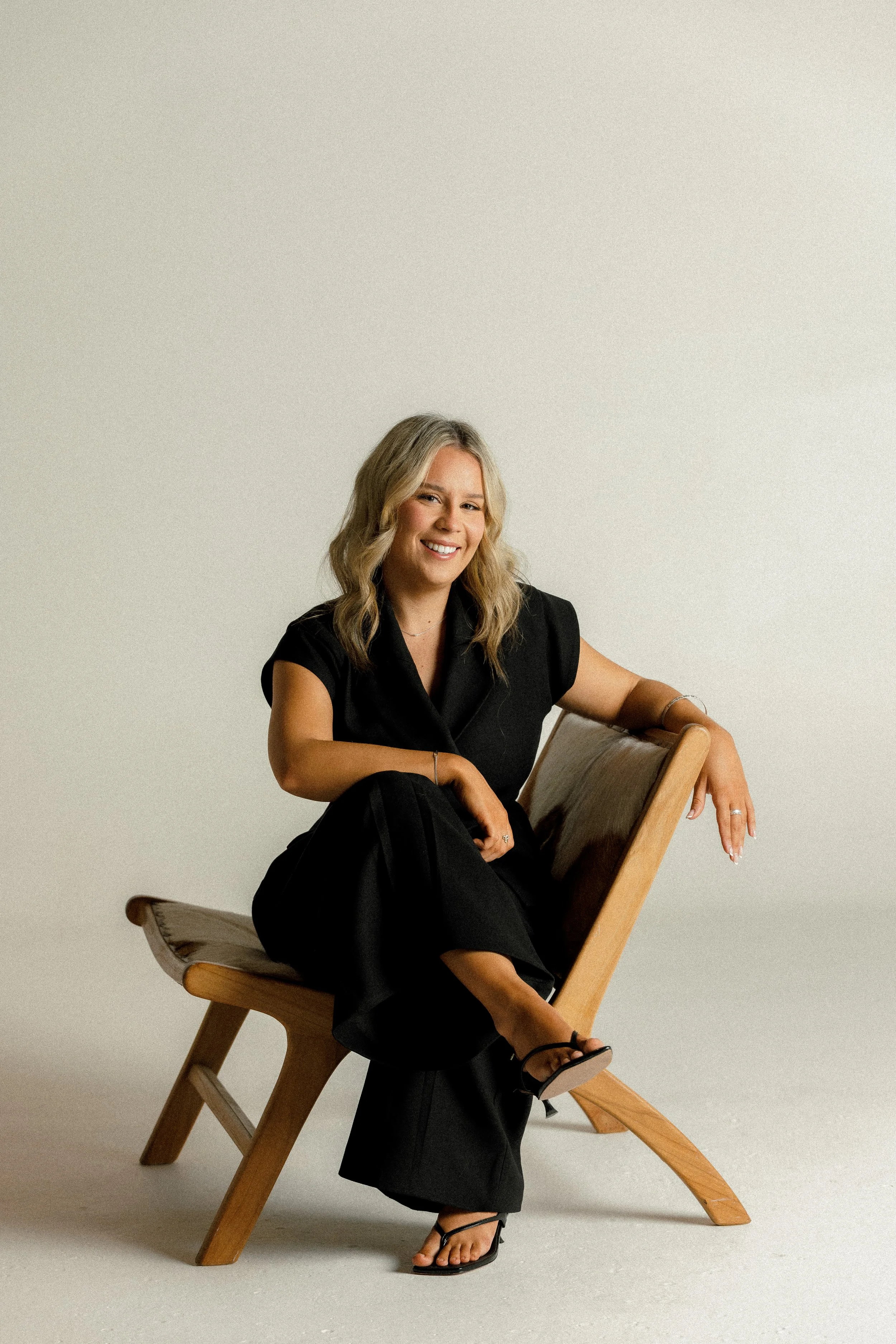 A woman sitting on a wooden chair, smiling, wearing a black outfit and black sandals, against a plain neutral background.