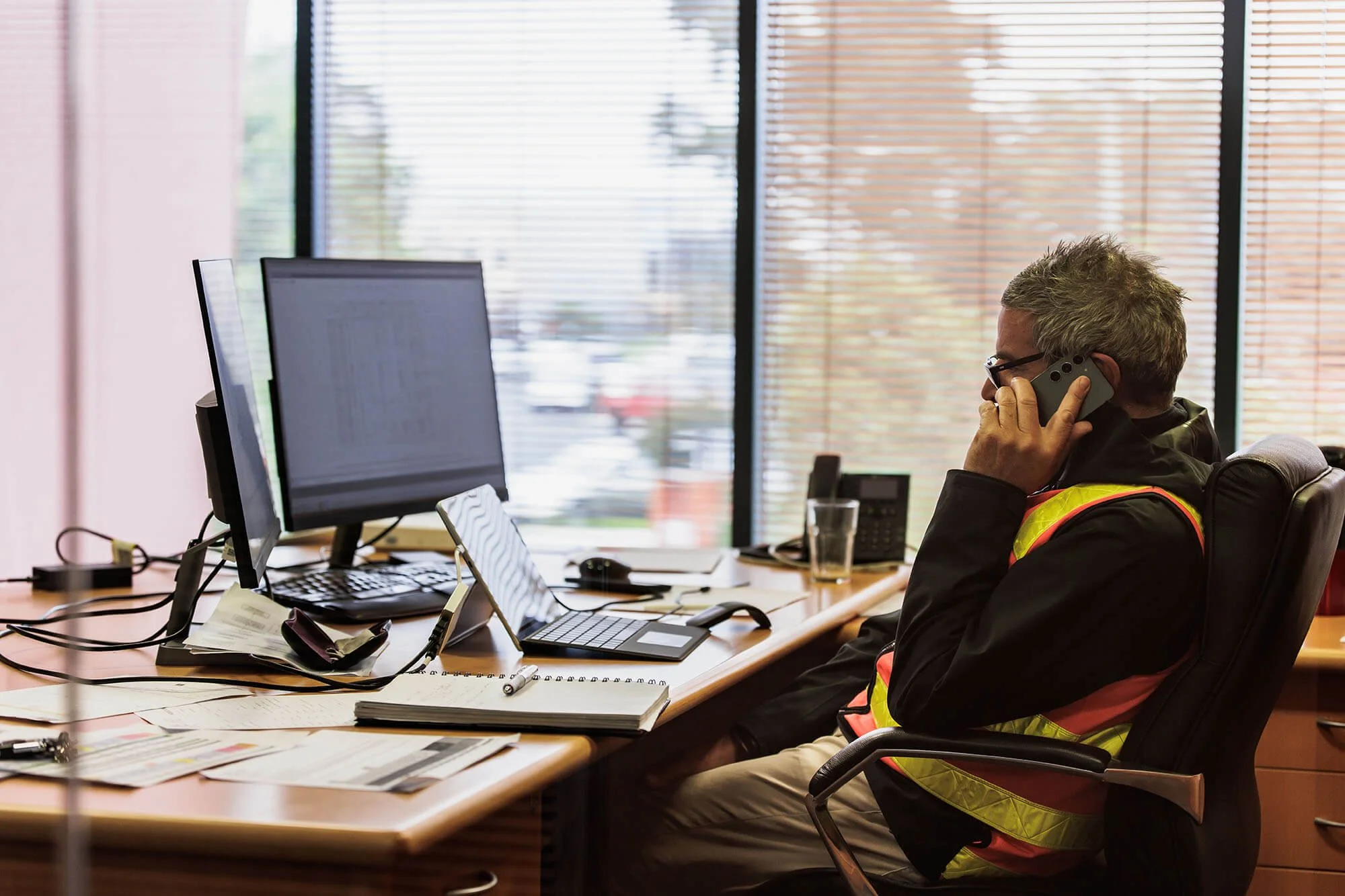 A man in hi-vis on the phone at a desk with a laptop in front of him.
